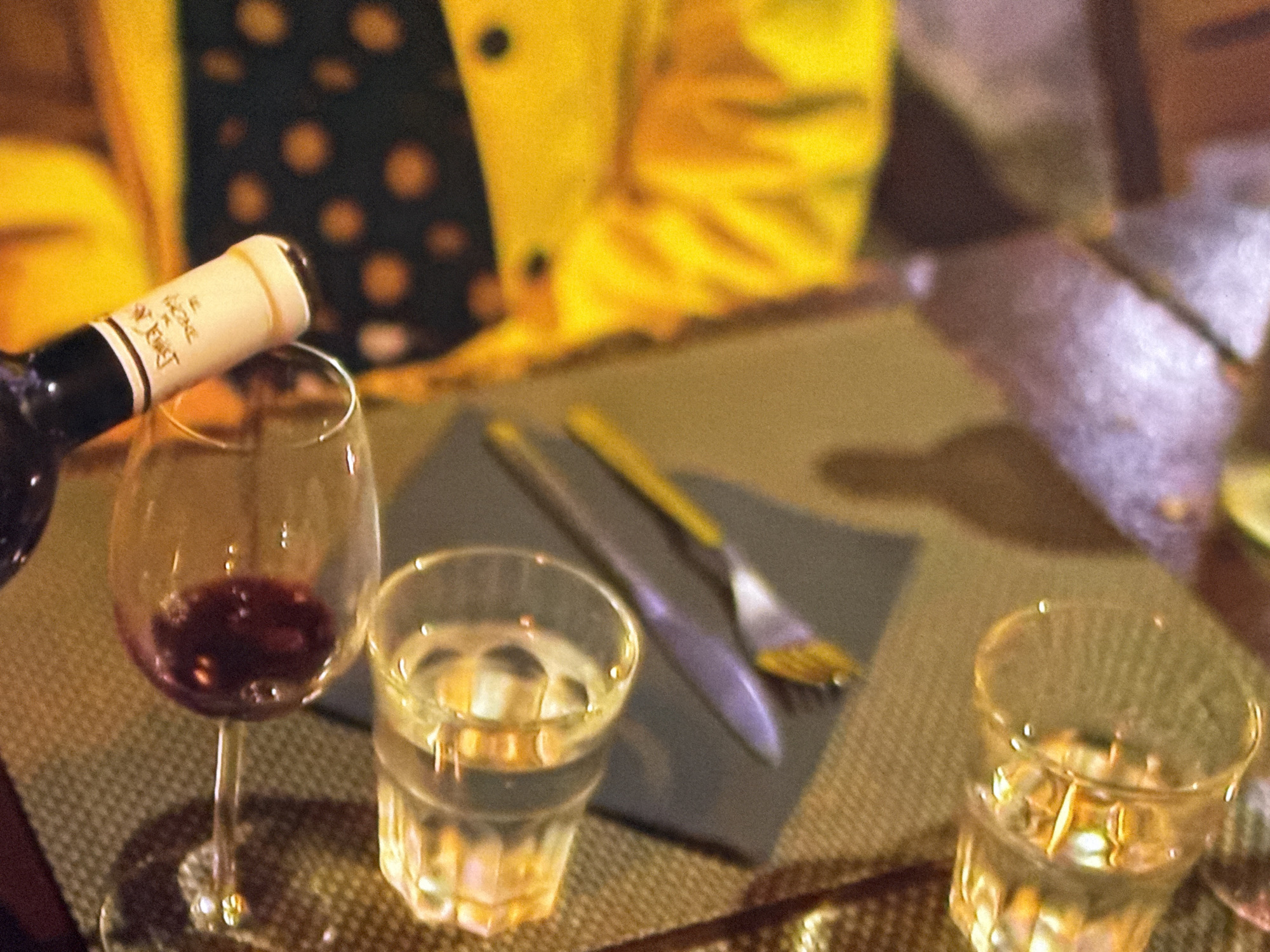 The image shows a table setting in what appears to be a restaurant or dining environment. A partially filled wine glass sits next to a glass of water, and a wine bottle is tilted as if pouring into the wine glass. The table is set with silverware, and a portion of a person wearing a yellow garment with brown polka dots is visible in the background. The exact location is not discernible from the image.