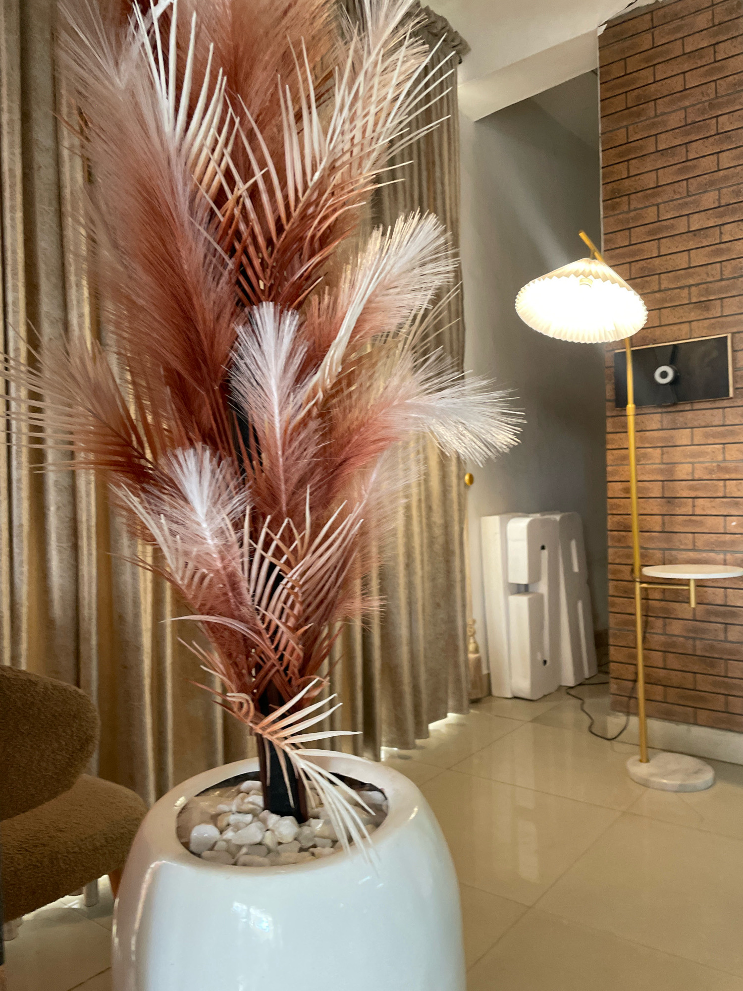 The image depicts an indoor scene featuring a tall, decorative plant with dried fronds in shades of terracotta and white, housed in a large, white, ovular planter. The planter is filled with white pebbles. To the left of the plant, a textured, brown upholstered armchair is partially visible. In the background, beige curtains hang behind the plant. Further to the right, a floor lamp with a pleated, white lampshade stands near a brick wall. The lamp has a thin, gold-colored pole and a round, marble base. A small, round shelf is attached to the lamp's pole. Mounted on the brick wall is a black rectangular object with a white circular element. A white, rectangular appliance, possibly an air conditioner or water dispenser, is positioned on the floor to the right of the curtains. The floor is tiled with large, light-colored square tiles that reflect the light. The scene appears to be set in a well-lit interior space, likely a living room or lounge area. The location is described as being in Ibadan, Nigeria.