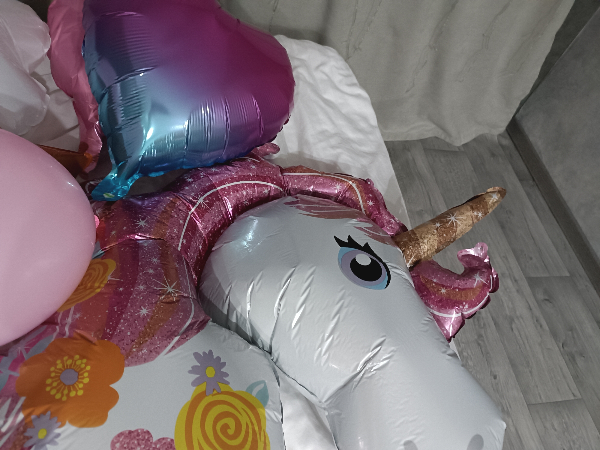 The scene depicts multiple deflated or partially inflated balloons resting on a bed and the adjacent floor within a room in Unknown, Unknown. Dominating the foreground is a white foil balloon shaped like a unicorn's head, featuring a black eye with a purple iris and light blue eyelid, and a brown, star-patterned horn. Partially covering this unicorn balloon is a deflated, gradient pink-to-blue heart-shaped foil balloon. A round, light pink latex balloon is visible on the far left. Other foil balloons, patterned with orange and purple flowers, yellow swirls, and pink backgrounds with white speckles, are visible beneath and around the primary balloons. The balloons rest on a rumpled white bedsheet, which is bordered by a light grey wooden plank floor on the right side of the image. In the background, a light-colored, vertically hanging, textured curtain is visible. No individuals are present.