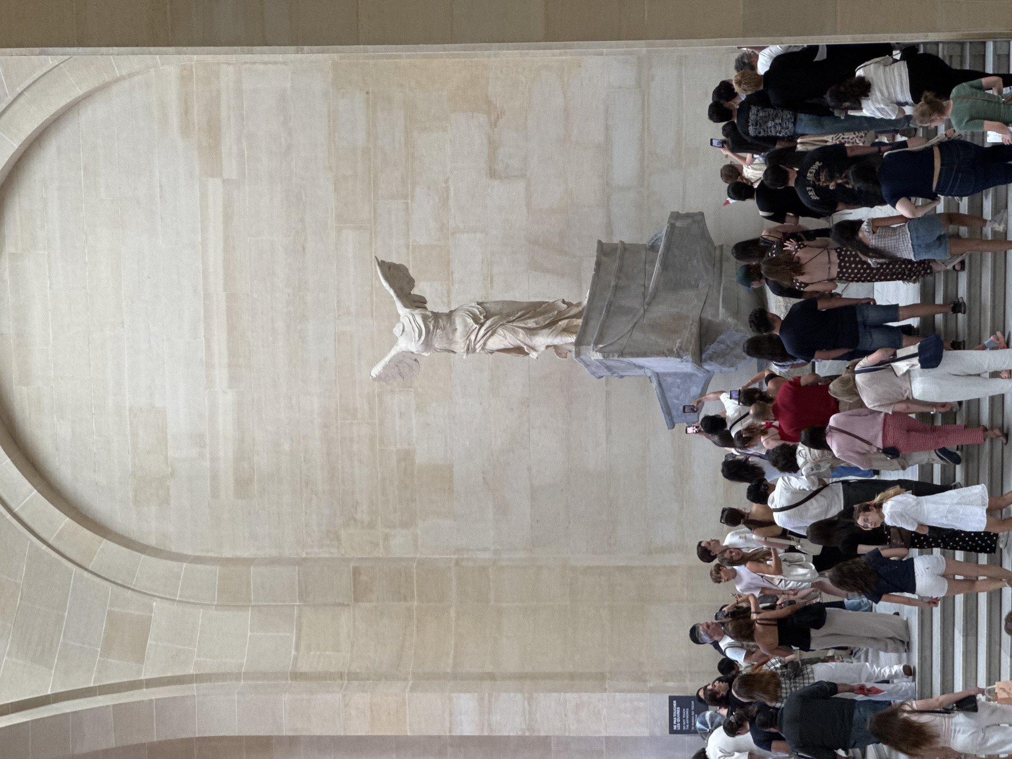 The image depicts the Winged Victory of Samothrace, a renowned ancient Greek sculpture, displayed in a grand hall with stone architecture. The sculpture, headless and with wings outstretched, stands on a stone pedestal. A crowd of tourists, dressed in casual modern clothing, are gathered around the base of the sculpture, many holding up smartphones to take pictures. The hall features large arched openings and walls constructed of light-colored stone blocks. A sign in French and English advising visitors not to touch the artworks is visible on the left. The scene suggests a cultural institution, likely the Louvre Museum in Paris, France, given the prominence of the Winged Victory.