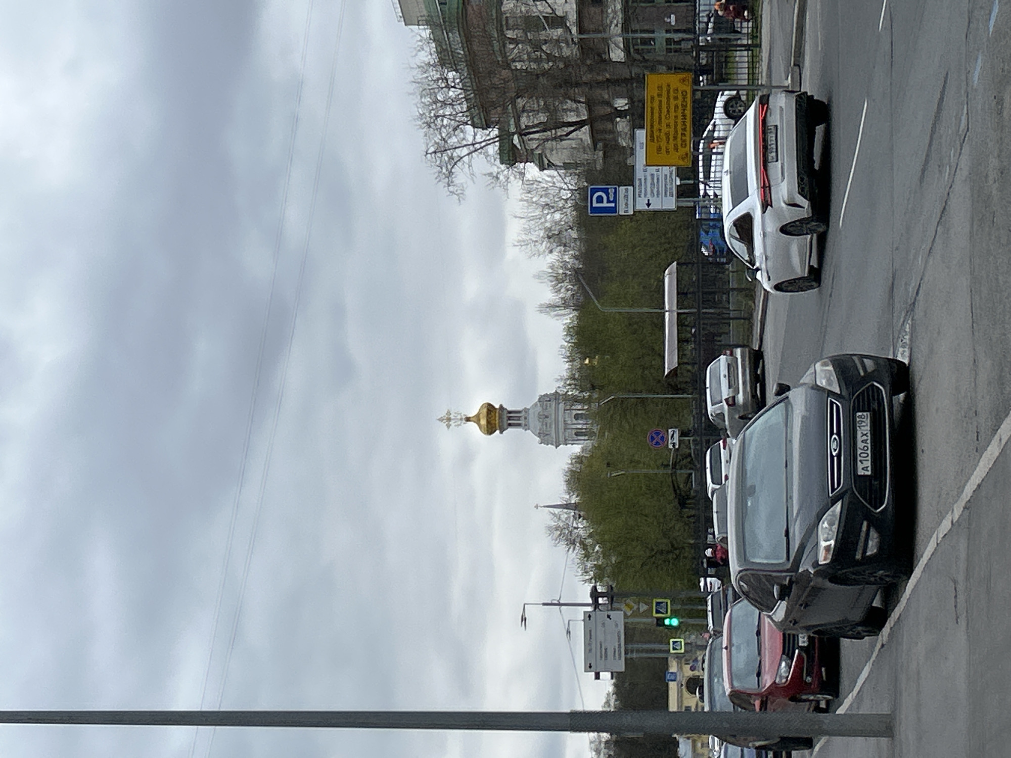 The image captures an urban street scene in Saint Petersburg, Russia, on an overcast day. The perspective is from a vehicle on a multi-lane road, looking towards the right where buildings, trees, and a church are situated.
In the foreground, a dark grey Ford Kuga/Escape SUV with Russian license plate "A 106 AX 198" is prominent. Behind it, a red car is partially visible. Further along the road, a white Lexus sedan with license plate "H 699 YT 178" is seen, indicating local registration from Saint Petersburg regions. Other vehicles are also partially visible, suggesting an active street or parking area.
On the right side of the street, a mix of architectural styles is visible, including older buildings with intricate details and green-tinted roofs. Trees with sparse foliage, some just beginning to bud with green leaves, line the street. Dominating the background is a majestic white Orthodox church or cathedral featuring a distinctive golden onion dome topped with a cross, partially obscured by the trees.
Several street signs are visible. A blue sign with a white "P" indicates parking. Two yellow rectangular signs display text in Russian; one clearly shows "ОГРАНИЧЕНО" (Restricted/Limited), and another mentions "ПЛАТНАЯ ПАРКОВКА" (Paid Parking) and "ОГРАНИЧЕНО", providing details about local traffic or parking regulations. A green traffic light is also visible in the distance. The sky is uniformly grey and cloudy, indicating an overcast daytime setting without direct sunlight. Thin power lines are faintly visible across the upper sky.