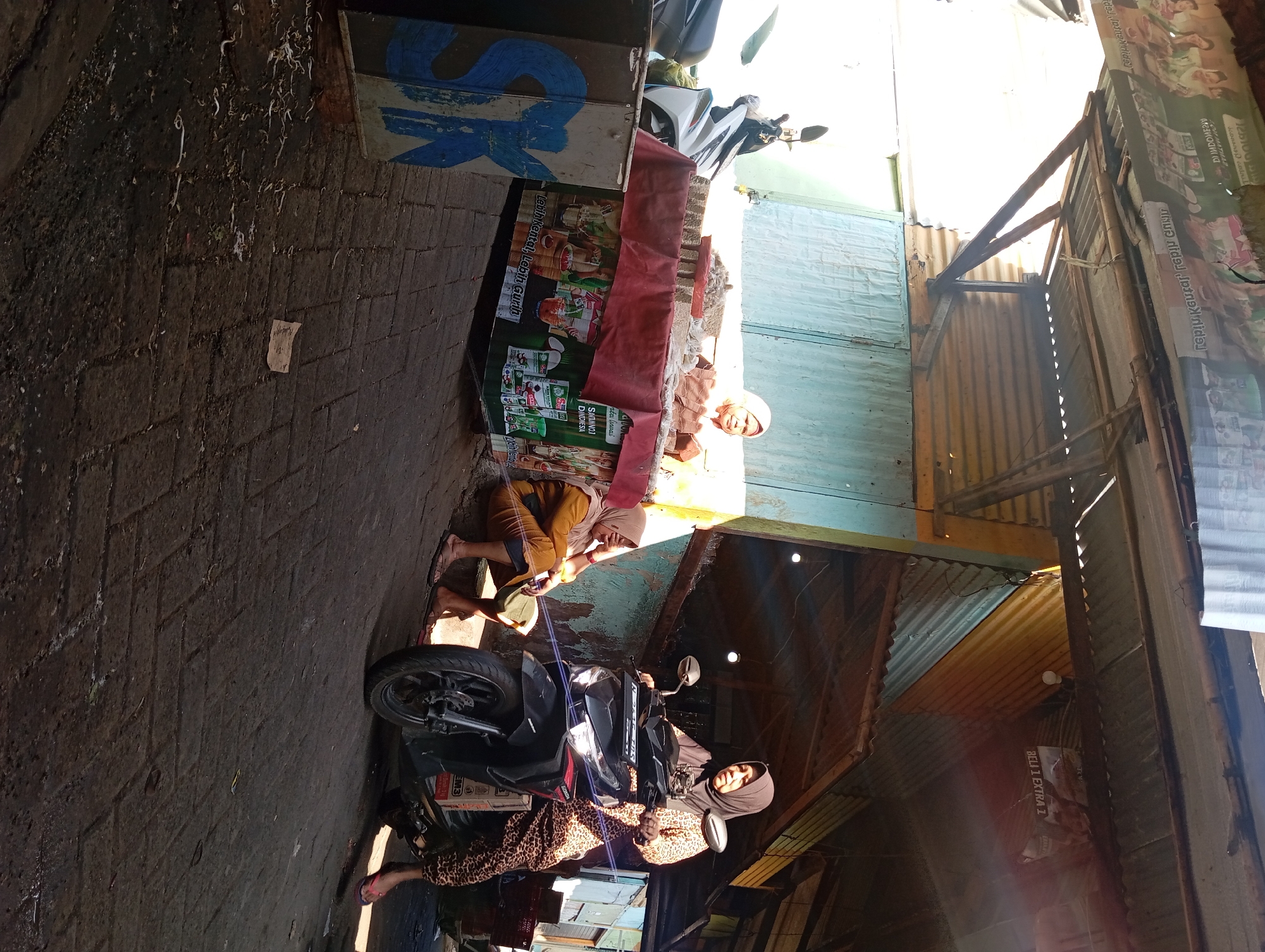 A woman wearing a headscarf and a leopard-print outfit is seated on a black Honda scooter at a market stall. The scooter is facing right, with its headlights illuminated. To the left of the scooter, another woman in a yellow headscarf and brown outfit is sitting on a step, holding a yellow object. She is looking down with her hand on her forehead. The market stall is covered with a red cloth and displays various packaged goods. A motorcycle is parked behind the stall. The ground is paved with brickwork. The scene appears to be in an outdoor market in Kepulungan, Indonesia, under a corrugated metal roof structure. A sign on the right side of the image reads "BELI 1 EXTRA 1".