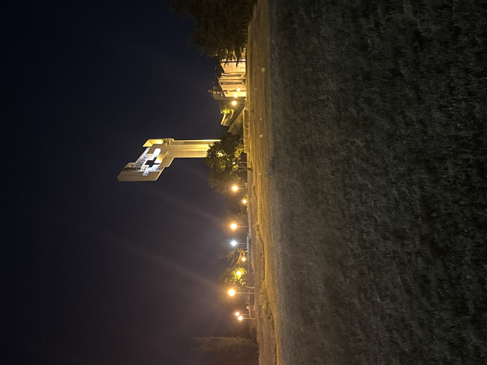 The image shows a nighttime view of the Defenders of Stara Zagora Memorial Complex in Stara Zagora, Bulgaria. The monument is illuminated and stands against a dark sky. The foreground contains grass and a path with streetlights leading towards the monument. The photo is taken at ground level, with the camera oriented vertically.