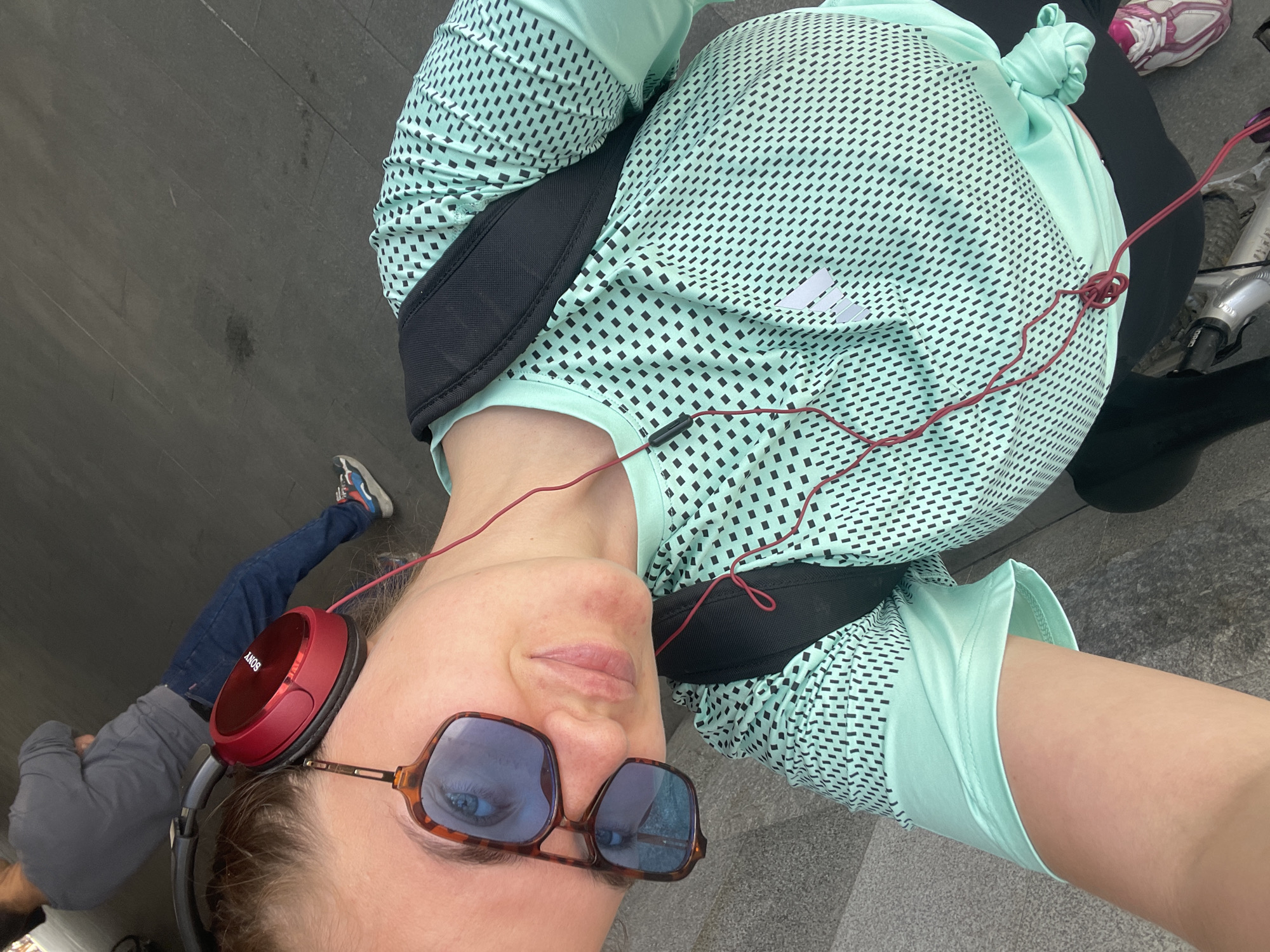 A woman takes a selfie outdoors, likely in a public space. She is wearing sunglasses, red over-ear headphones, and a light green athletic shirt with a black pattern, as well as black athletic shorts. A black strap or belt is visible over her shoulder. In the background, other people are walking, and a bicycle is visible. The ground is made of gray stone tiles. It cannot be definitively verified if the location is in Moscow, Russia without additional information.