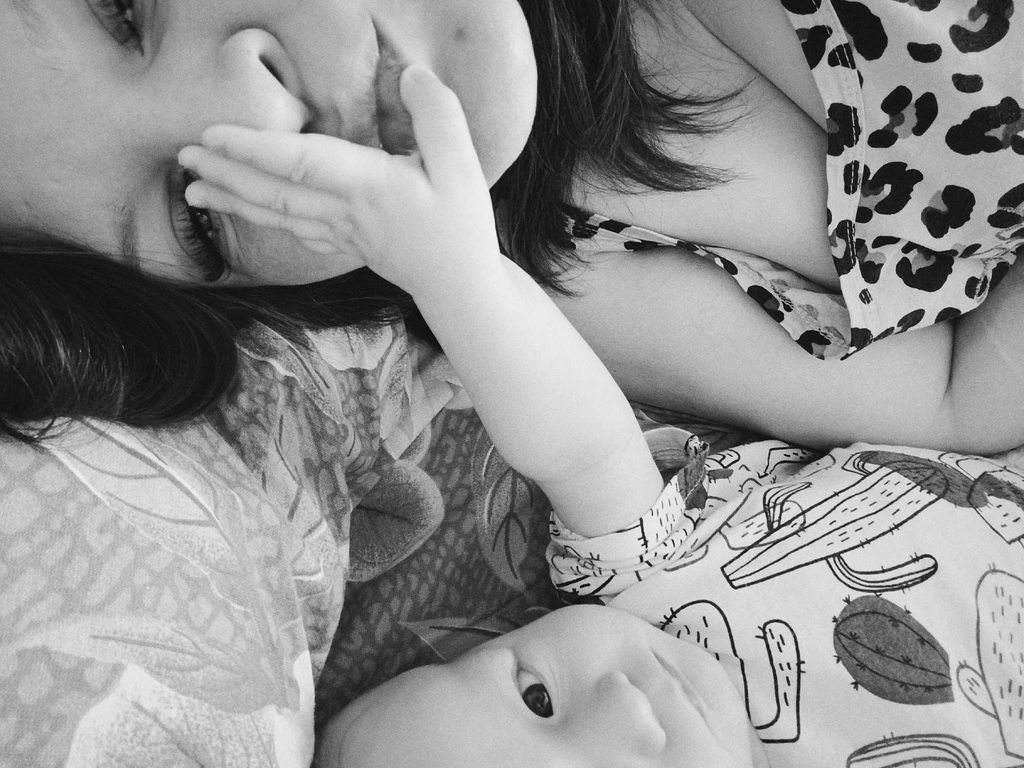 A black and white close-up shows a woman and baby lying down. The woman is on her side with her face turned towards the camera, her hand partially covering her mouth. She has dark hair. She is wearing a floral patterned shirt. Next to her is a baby lying on its back, facing the same direction as the woman. The baby is wearing a shirt with a cactus pattern. The location is not discernible from the image. There is no indication the photo was taken in Tobolsk, Russia.