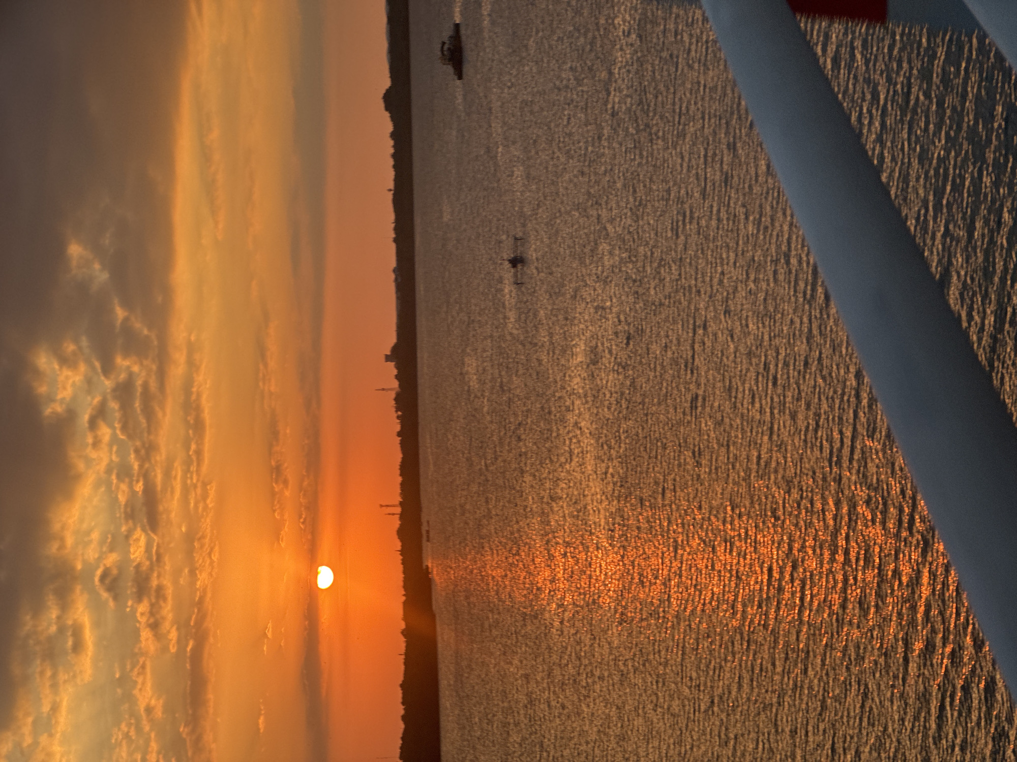The image captures a sunset over a body of water, likely the ocean, in Kuta Selatan, Indonesia. The sky is a vibrant orange and yellow, filled with scattered clouds. The sun, a bright disk, is low on the horizon, casting a shimmering reflection on the water. In the distance, a silhouetted coastline with trees and some structures is visible. There are two small boats on the water, one closer to the left and another further to the right. The foreground shows a part of a railing, suggesting the vantage point is from a boat or a pier. The water appears calm with ripples on its surface, reflecting the warm hues of the sunset.