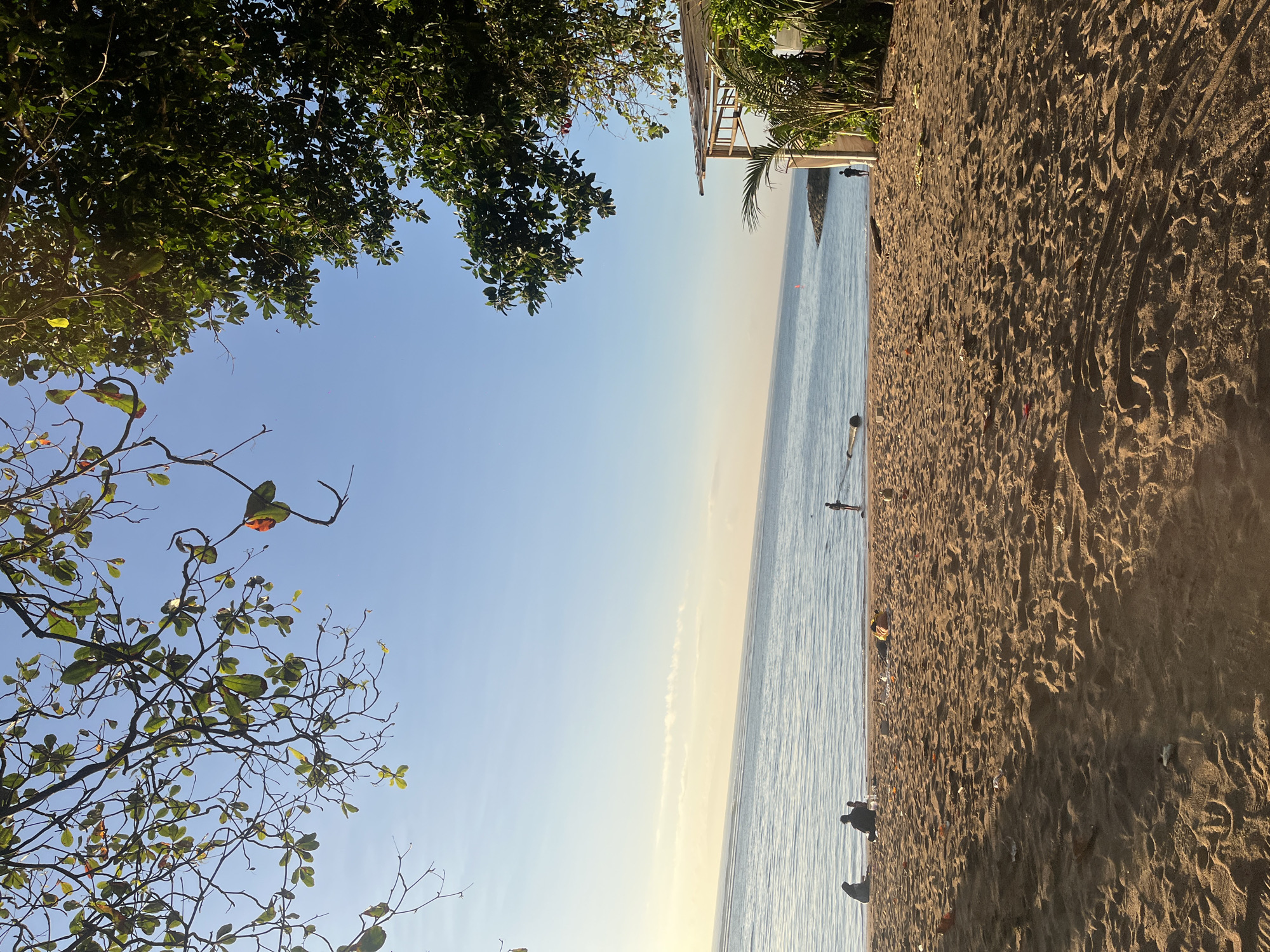 This is a ground-level shot of a beach scene at Denpasar, Indonesia. The foreground is dominated by sand with scattered debris. In the mid-ground, a calm body of water meets the sandy shore, with three people sitting on the beach near the water's edge and one person standing in the water. The background shows a clear blue sky transitioning to a lighter hue near the horizon, with a faint cloud streak. Lush green trees frame the top and right sides of the image, and a simple open-air structure with a thatched roof is visible on the right. The overall lighting suggests it is either early morning or late afternoon.