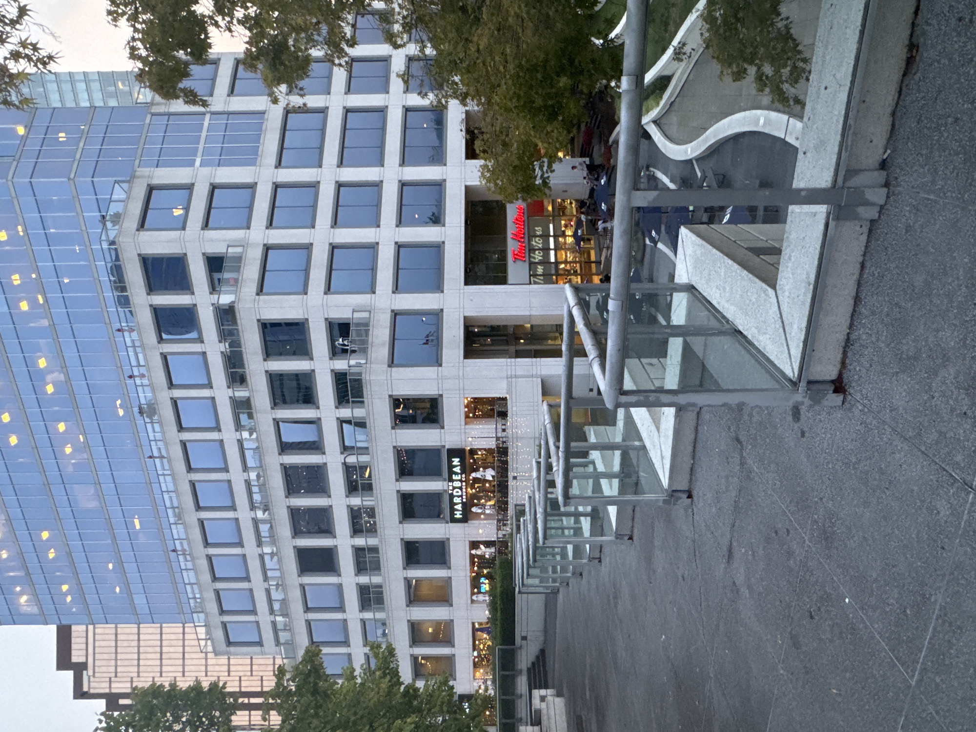 The image depicts a modern, multi-story commercial building in Vancouver, Canada. The building features extensive glass facades and a stone-clad lower section. Prominently visible on the ground floor are two commercial establishments: "The Hard Bean Brunch Co." and "Tim Hortons." Outside, there are tables and chairs for outdoor seating, some covered by umbrellas. A paved walkway with glass railings leads the viewer's eye towards the building and outdoor seating areas. Green trees with foliage are visible on the right side of the image, suggesting an urban park or landscaped area. The sky is overcast, indicating a cloudy day.