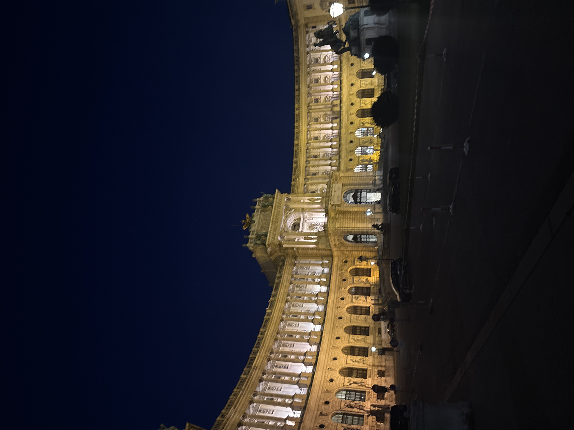 The image shows a night view of a grand, ornate building with many windows, illuminated by warm artificial lights. The building has a classical architectural style with columns and detailed facades. To the right of the building, there is a dark street with a few parked cars and some streetlights. A dark, starless sky forms the background on the left side of the image. The perspective is from a low angle, looking up towards the building. The overall scene appears to be in a city, likely a public space or a prominent landmark, given the scale and design of the architecture. There is no visible text in the image. The time of day is clearly night, and the weather appears to be clear. The mood is somewhat imposing due to the grand architecture and the darkness.