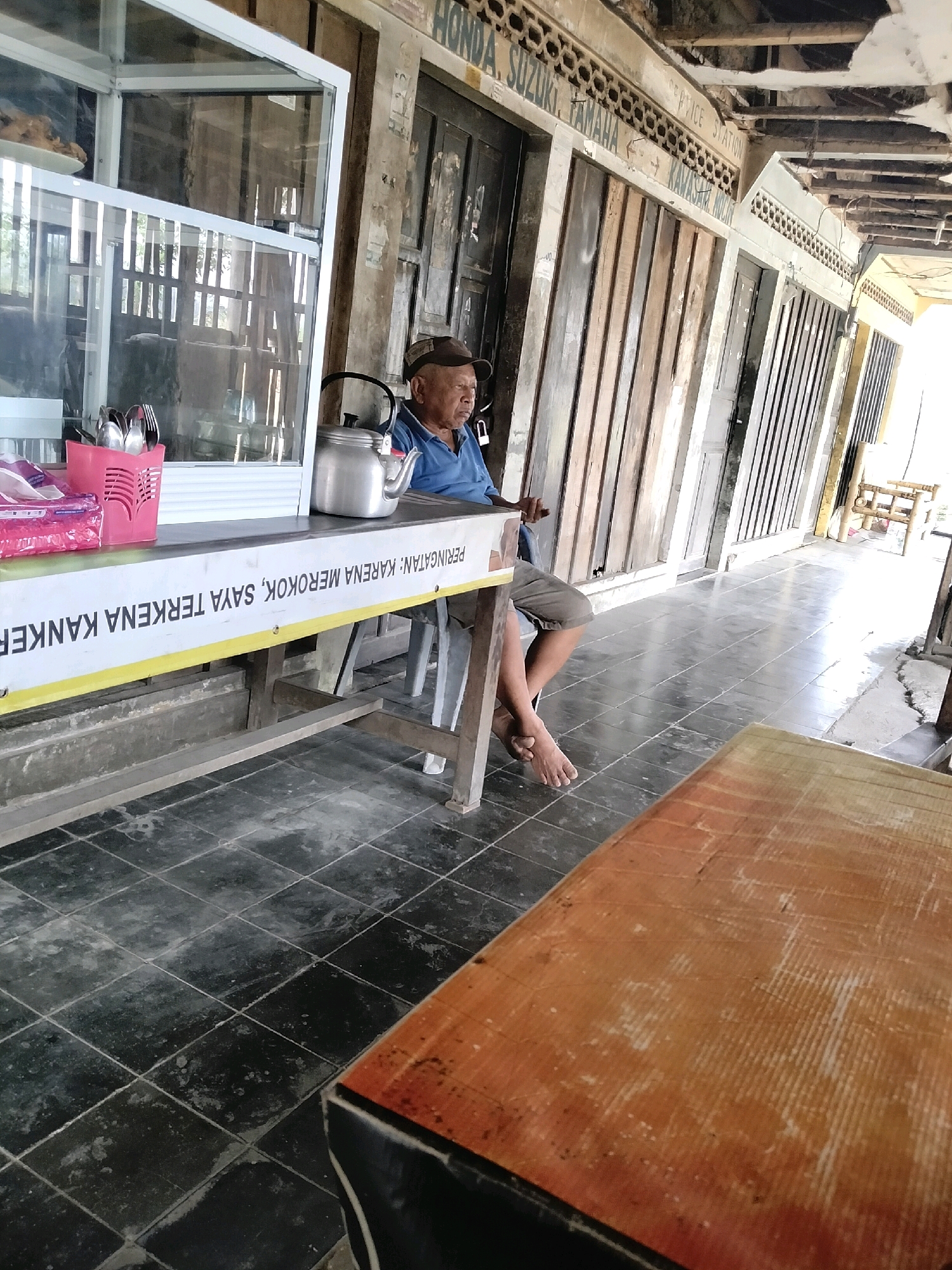 The image depicts an outdoor scene with an elderly man sitting at a table. The man is wearing a blue shirt and a dark cap, and is seated on a chair to the right of the frame. In front of him is a table with a silver kettle and a pink basket holding cutlery. A banner with text is draped across the table. Behind the man, there are several wooden doors and an overhead structure that appears to be part of a shop or market. The ground is tiled. To the left of the man is a glass display case containing food. In the foreground, a portion of another table, made of wood with an orange finish, is visible. The overall impression is that of a casual outdoor dining or resting area.