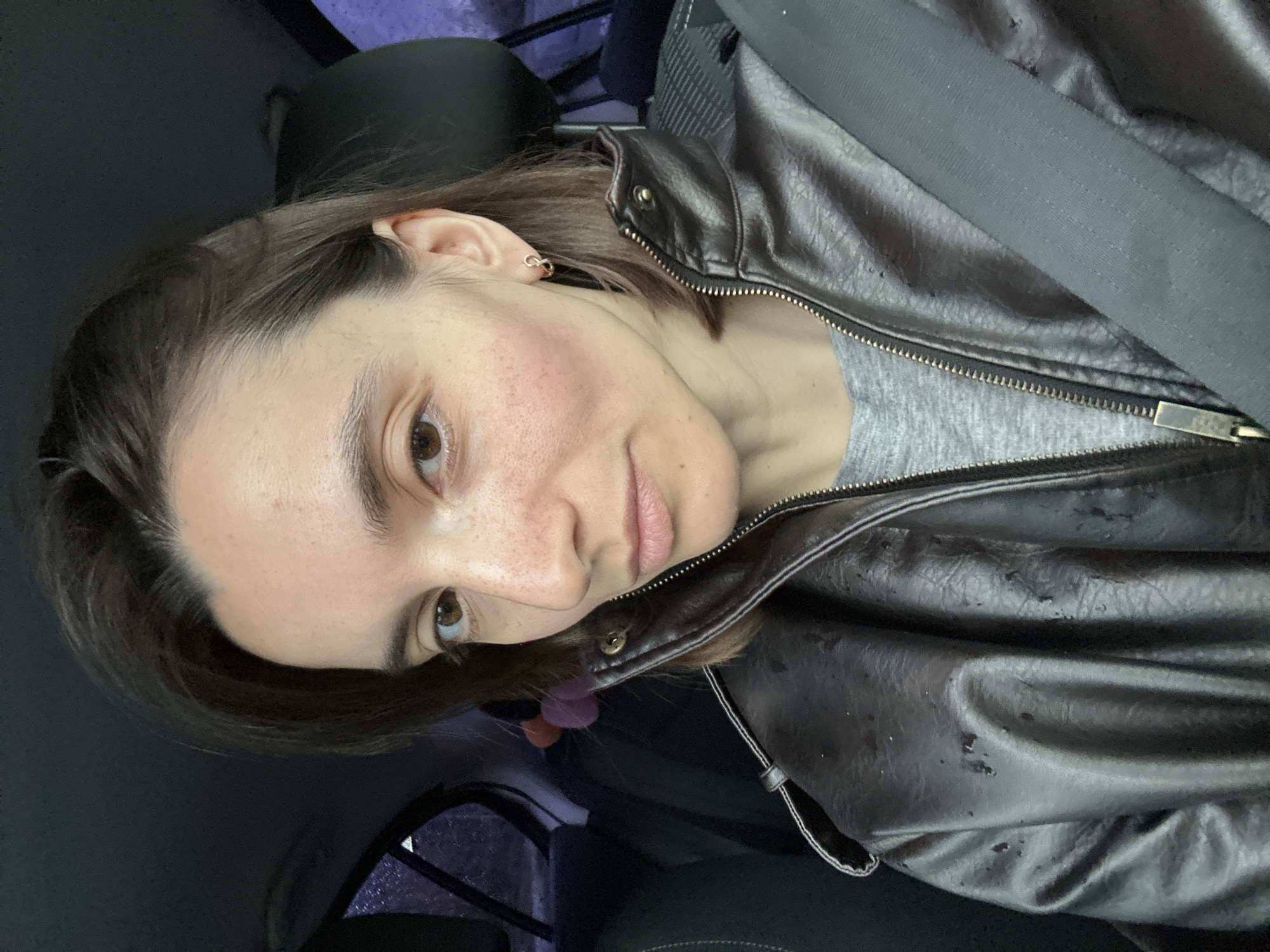 A woman is in a car, wearing a black leather jacket and a grey shirt. She is wearing a seatbelt. The background suggests an urban environment. It is not possible to verify the location as Moscow, Russia from the image alone.