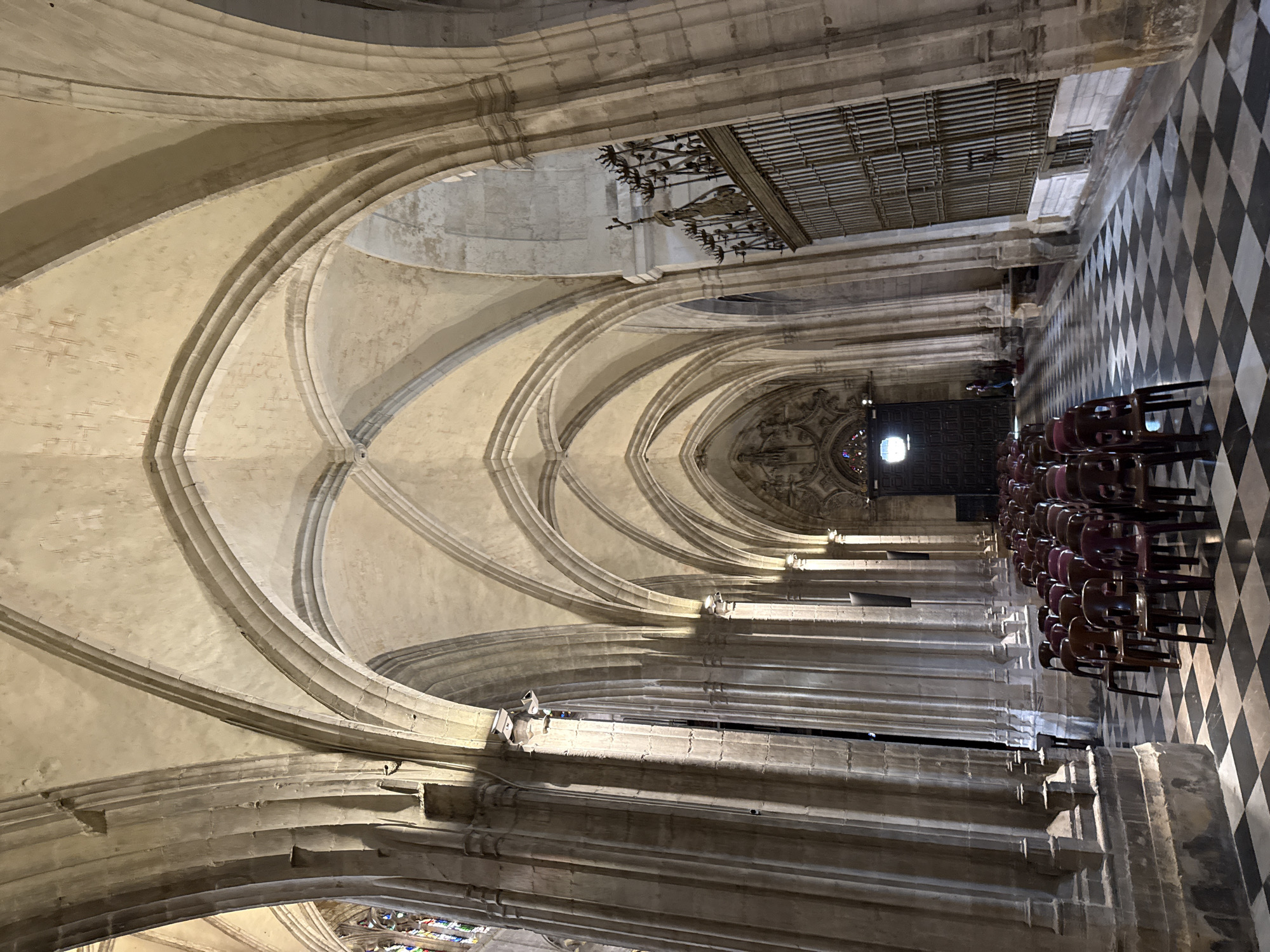 A photograph depicts the interior of a cathedral or similar large religious structure. The scene includes high, arched ceilings constructed of stone, supported by columns that run down the side of a long aisle. On the right side of the aisle, rows of chairs are arranged on a floor tiled with a black and white checkered pattern. At the end of the aisle is a large wooden door, possibly an entrance. The environment appears to be dimly lit, with light filtering in from windows. There are no people visible in the photograph. The location is Unknown, Unknown.