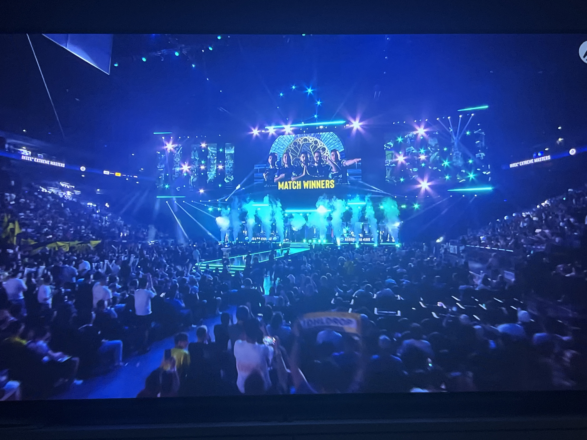 The image depicts a large-scale esports event, likely Intel Extreme Masters, taking place in an indoor arena. A stage is set up with bright blue and green lighting, accompanied by pyrotechnics or smoke effects. On the stage, a group of individuals are posing for a photograph under a large screen displaying the text "MATCH WINNERS" and a decorative arch. The audience, filling the arena seats, is visible in the foreground and throughout the venue, many holding up their phones or arms. The overall atmosphere is celebratory and energetic, characteristic of a major competitive gaming event. The location in Varna, Bulgaria, is not visually evident from the image itself.