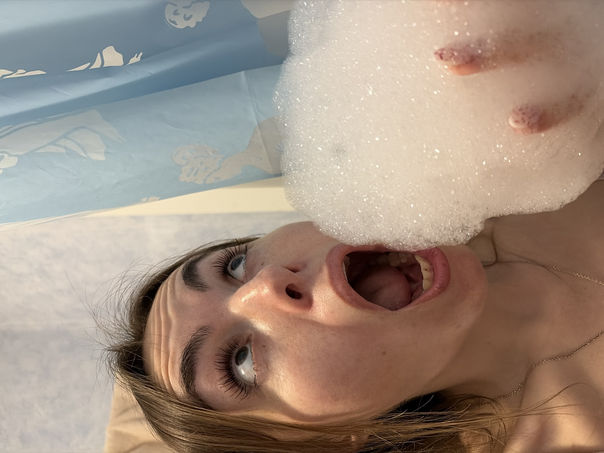 A low-angle shot depicts a woman positioned in a bathtub. A mass of white foam, possibly bath bubbles, hovers directly above her open mouth. Her eyes are directed upwards, away from the foam. She is inside what appears to be a bathroom with a blue shower curtain. The location in the city of Izhevsk, Russia cannot be determined from the image.