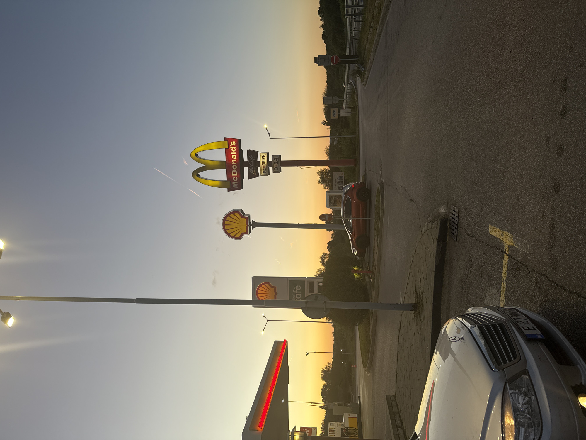 The image depicts a roadside scene, likely a gas station or rest stop area, near Studena, Bulgaria, at what appears to be dusk or dawn. Dominating the view are illuminated signs for McDonald's and Shell, indicating the presence of these establishments. A gas station is visible. Several cars are parked at the location. The lighting suggests it is either early morning or late evening. The sky fades from a light blue near the top to a warm orange/yellow near the horizon. A road stretches into the distance. A silver car is parked in the foreground. No people are explicitly visible.
