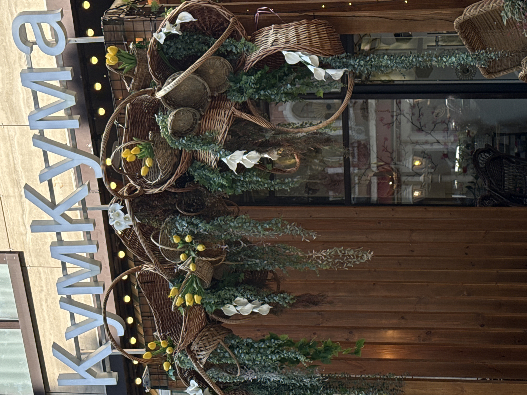 The image shows a street scene in Sochi, Russia. The Cyrillic word "КУЛИНАРИЯ" is written vertically in large, white block letters on the left side of the image. The letters are attached to a structure that is also lit by small, round lights. To the right of the signage is an arrangement of wicker baskets and various green plants with small white and yellow flowers. The baskets are mounted on the exterior wall of a building with horizontal brown wooden planks. Through the window of the building, an interior space is visible with decorative elements and what appears to be other pieces of furniture or decor.