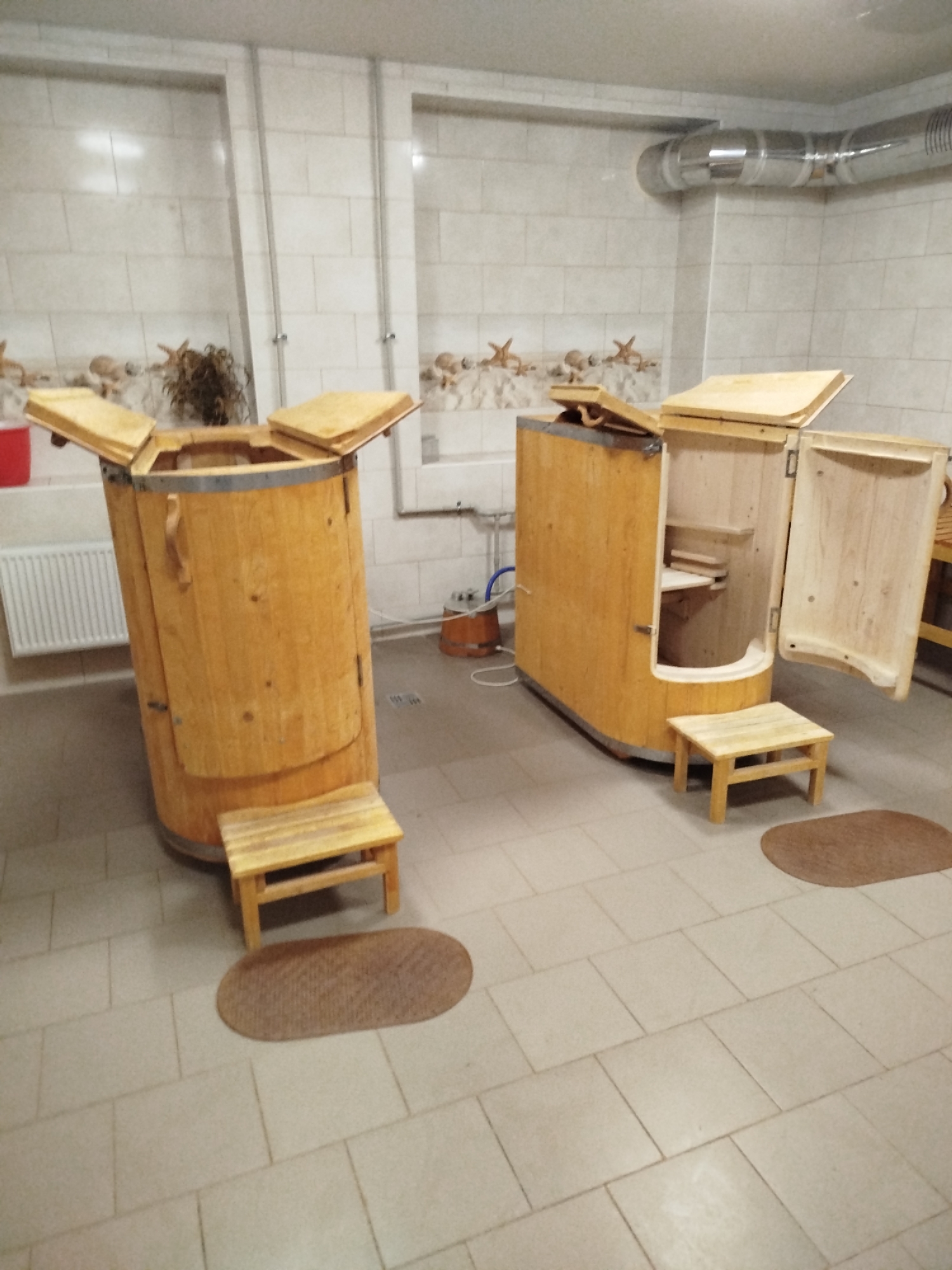 Two wooden barrel-like saunas are in a room with tiled floors and walls. The sauna on the left is a tall, cylindrical barrel with a hinged lid that is partially open. A small wooden stool is placed in front of it. The sauna on the right is a longer, more rectangular barrel shape with a door that is open, revealing an interior shelf. A small wooden stool is placed in front of it as well, with a mat to its right. Both saunas appear to be made of light-colored wood and have metal banding around them. In the background, a white radiator is visible on the left wall. There is also a metal ventilation pipe running along the ceiling. The walls are tiled with a light-colored pattern. No people are present in the image.