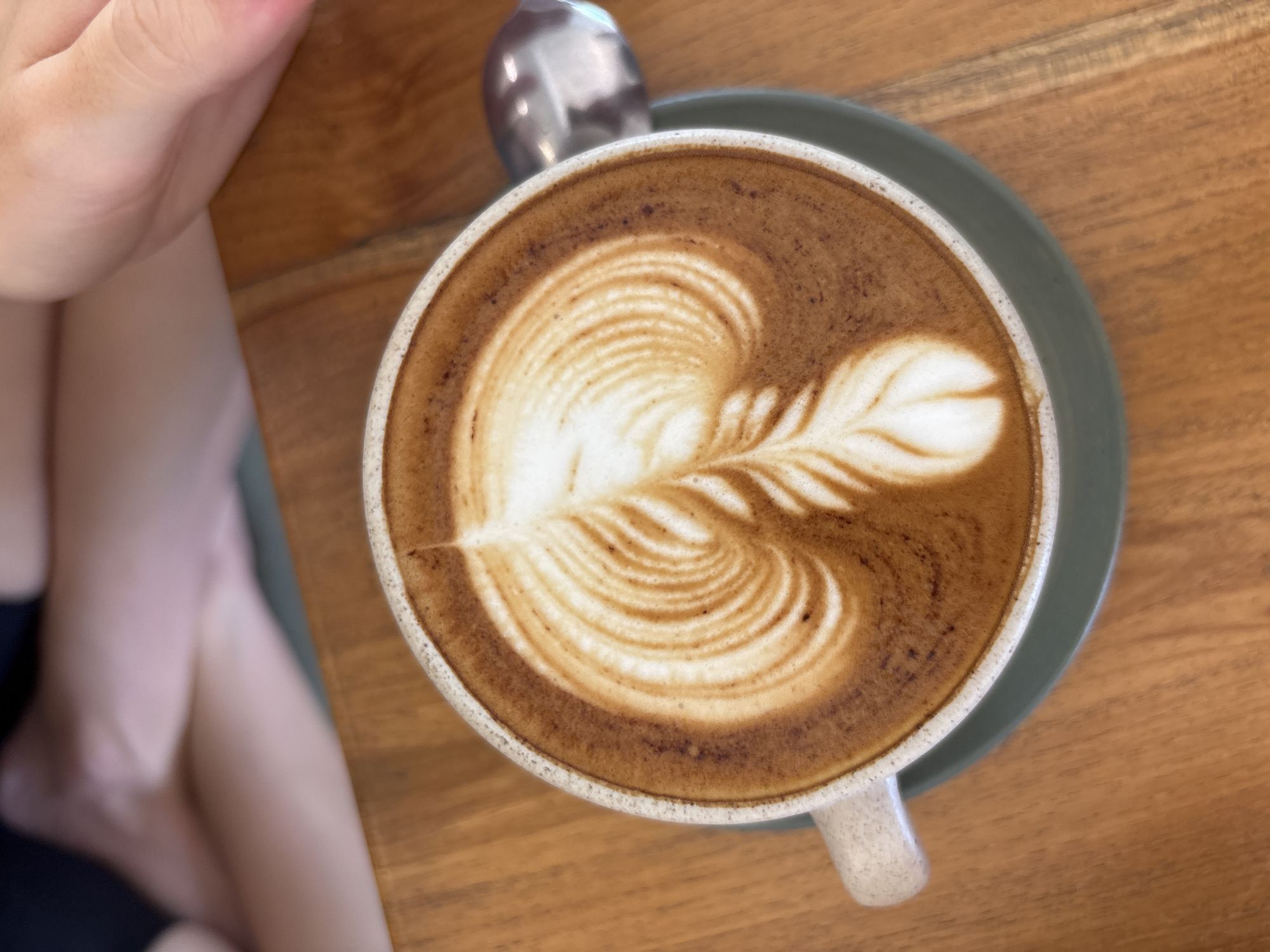 Overhead shot of a cappuccino with latte art in a white ceramic mug on a grey saucer, placed on a light brown wooden table. A spoon rests near the top of the saucer. Partial view of a person's leg is visible on the left side of the frame. The location is speculated to be in Kuta Selatan, Indonesia.