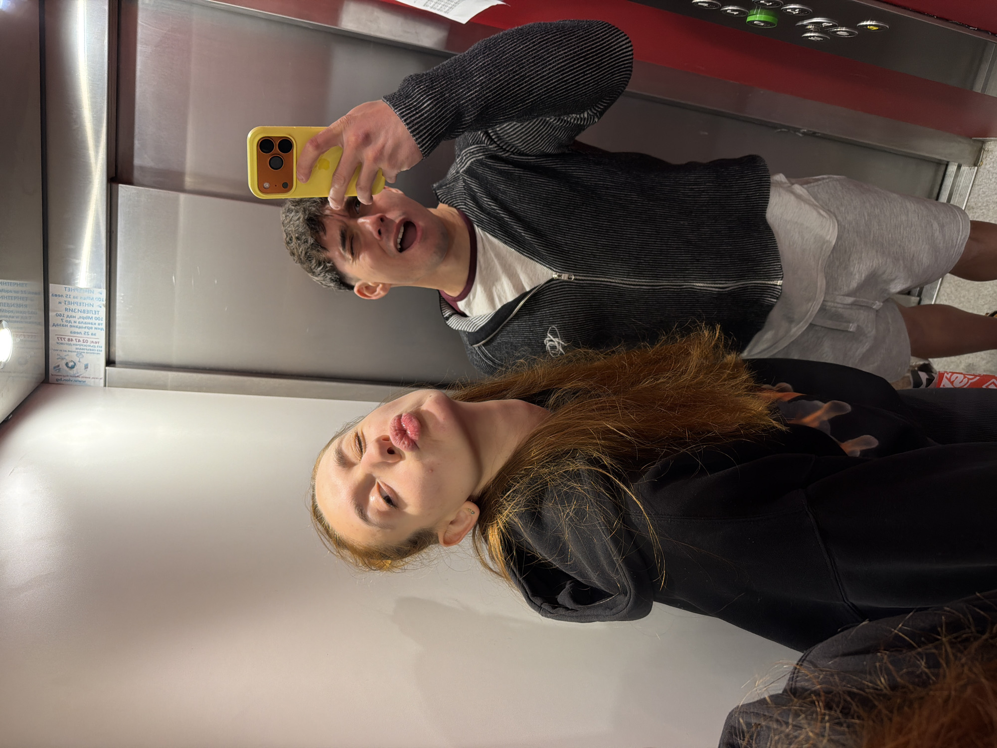 The image depicts a selfie taken in an elevator. Two individuals are visible: a young woman in the foreground with long auburn hair, wearing a black hoodie and a person in the background wearing a gray striped zip-up jacket, a white t-shirt and grey shorts, and holding a yellow smartphone. The interior of the elevator is made of stainless steel panels, with a row of buttons visible at the top. There are informational stickers attached to the side wall of the elevator. The image was taken at night in the city of Sofia, Bulgaria.