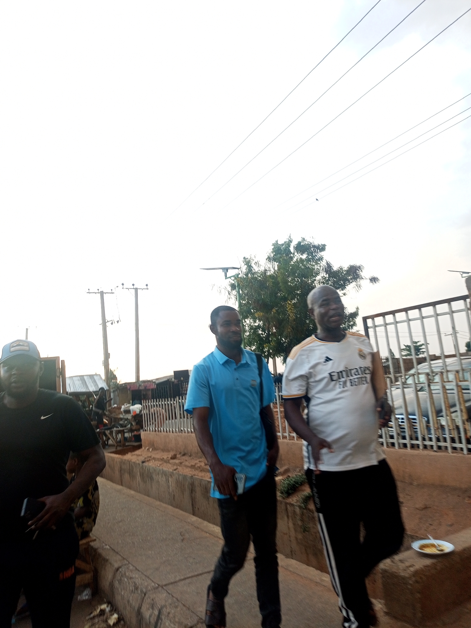 The image shows three men walking along a sidewalk in what appears to be a residential or commercial area in Gombe, Nigeria. The first man, on the left, is wearing a black t-shirt, black pants, and a baseball cap. The second man is wearing a blue short-sleeved shirt and black pants. The third man is wearing a white Real Madrid jersey and black track pants with a white stripe. They are all walking in the same direction.
In the background, there are utility poles, a tree, a metal fence, buildings, and parked vehicles. On the sidewalk, near the third man, there is a plate of food. The sky is overcast.