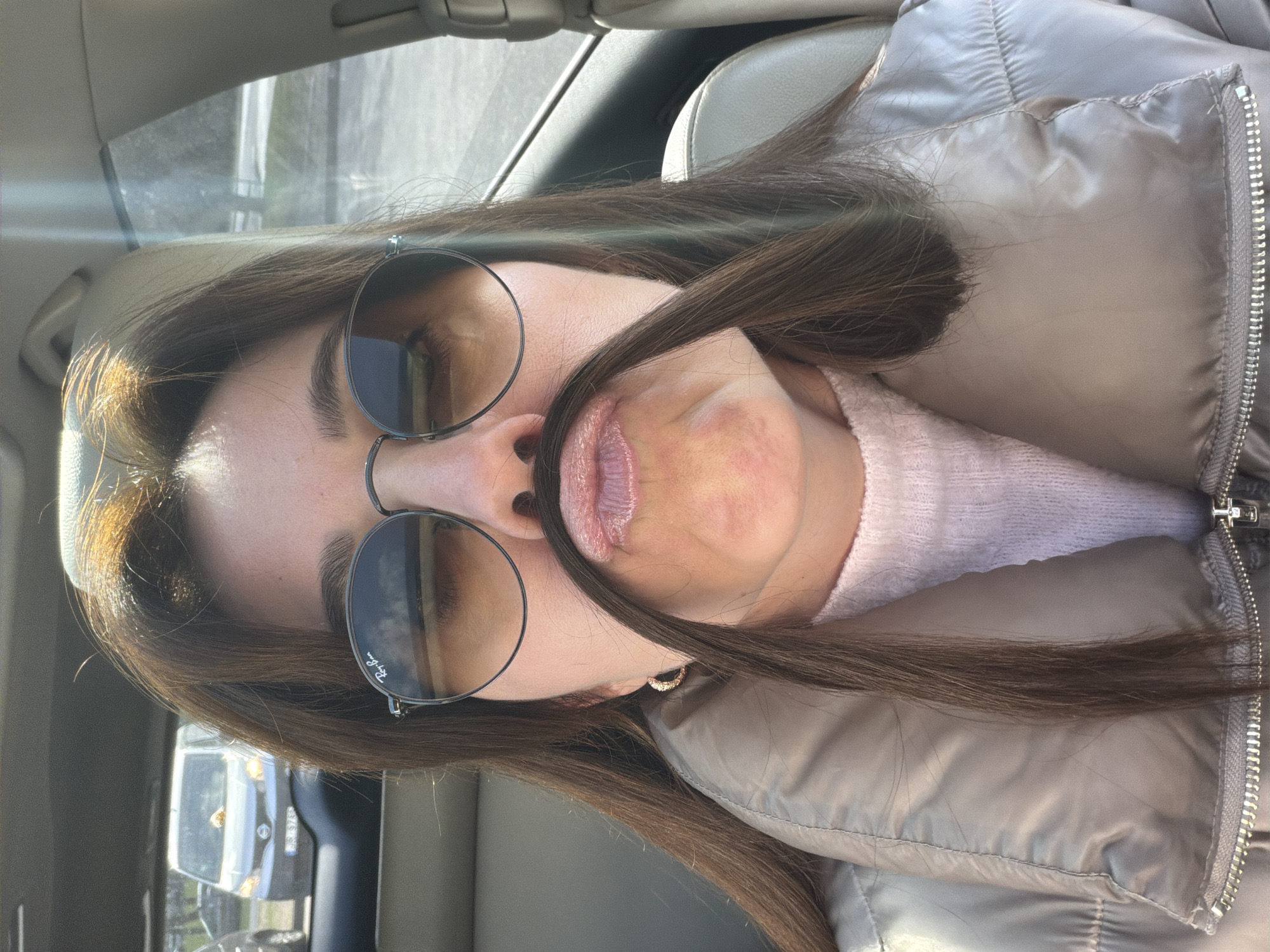 The image, oriented sideways, is a casual portrait of a young person, likely female, taken inside a car, possibly in Riga, Latvia. The individual has long, straight dark brown hair, which is playfully styled to create a 'mustache' over their lightly glossed, pinkish lips. They are wearing dark, round-lensed sunglasses with thin metal frames, bearing the 'Ray-Ban' logo on the lower right portion of the left lens. Their attire includes a light pink or peach-colored knitted sweater, visible at the neckline, worn beneath a quilted grey or tan puffer jacket with a prominent silver zipper running down the front. A small, decorative hoop earring with tiny stones is visible on their right ear.
The setting is a car interior, with parts of a light-colored headliner, a sun visor, and a car seat visible in the background. Bright natural light, likely sunlight, streams in from the left, illuminating parts of the subject's hair and the car's interior, suggesting a sunny daytime. The background through the car windows is out of focus, showing indistinct outdoor elements. The playful arrangement of the hair over the lips suggests a lighthearted and candid moment. No other specific visual cues indicating the city of Riga are present within the image itself, though the location context was provided.