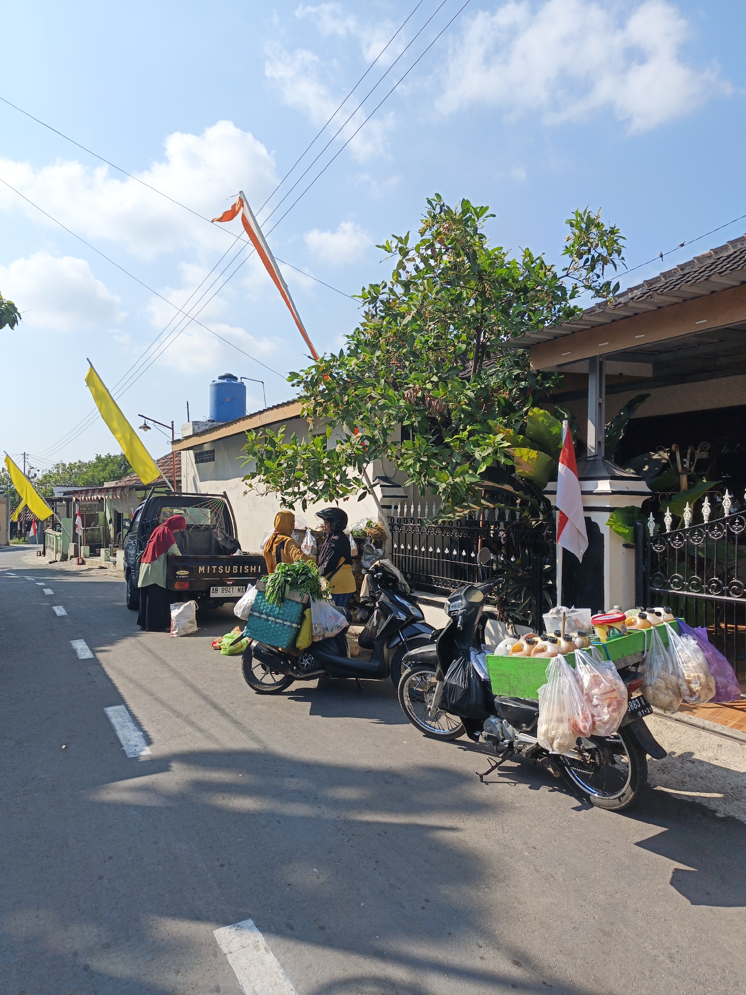 In Jatinom, Indonesia, on a sunny day, a street scene unfolds. A black Mitsubishi pickup truck is parked on the side of an asphalt road, with its tailgate down. Three individuals are near the truck; one in a red headscarf and green top, and two others in yellow and dark clothing. Bags of produce, including green vegetables, are visible. In front of the truck, a motorcycle with a green crate attached to its rear is parked. This crate is filled with various items, including bottles of liquid, containers, and bags of food products, suggesting it's a mobile food stall. Another motorcycle is parked beside it. The street is lined with houses and fences, and yellow and orange flags are flying from poles, adding a festive touch to the scene. The sky is blue with scattered white clouds.