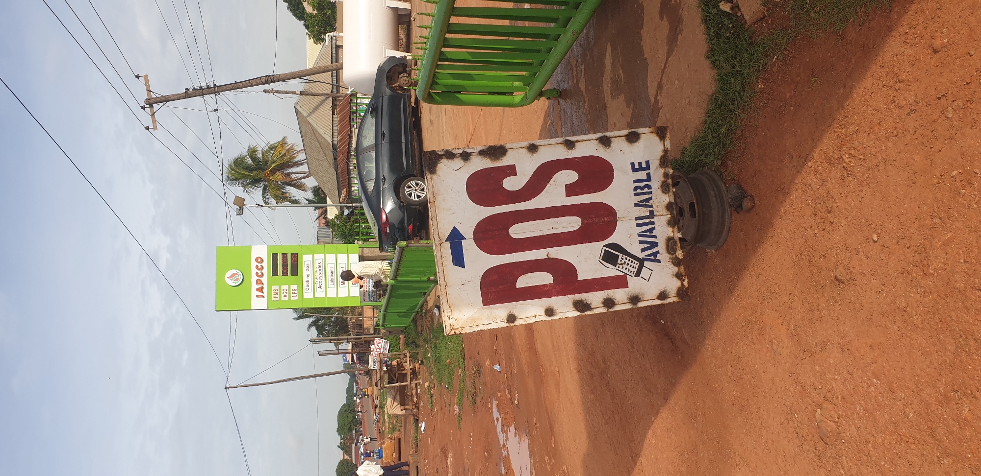 The image depicts an unpaved, reddish-brown dirt roadside scene in the city of Yelwa, Nigeria, under an overcast sky with patches of brighter light. In the immediate foreground, a weathered, rectangular white metal sign, propped up by a dark metal wheel rim, displays "POS" in large red letters with a blue arrow pointing right above it. Below, an illustration of a mobile POS terminal is visible, followed by the word "AVAILABLE" in blue text, indicating Point of Sale services.
Behind the foreground sign and to the right, a bright green metal barrier defines the perimeter of a JAPCCO gas station. A tall, green and white JAPCCO sign stands prominently, displaying "PMS", "AGO", and "LPG" with unreadable digital prices, followed by "Cooking Gas", "Cylinders", "Accessories", "Lubricants", and a partially visible "Market". A man in a light-colored long-sleeved shirt leans against the green barrier near the JAPCCO sign.
To the right of the man and the sign, a dark-colored sedan, possibly a Hyundai Elantra, is parked. The vehicle's front driver's side wheel is visibly missing, resting directly on its brake rotor. Further into the gas station area, large white cylindrical storage tanks are visible.
The ground across the scene is unpaved, consisting of reddish-brown dirt with several muddy puddles, suggesting recent precipitation. Patches of green grass grow alongside the barriers. In the mid-ground, a busy street is visible with other individuals, indistinct due to distance, and makeshift structures. Utility poles with numerous overhead wires traverse the upper half of the image. Palm trees are scattered throughout the background, indicative of a tropical climate.