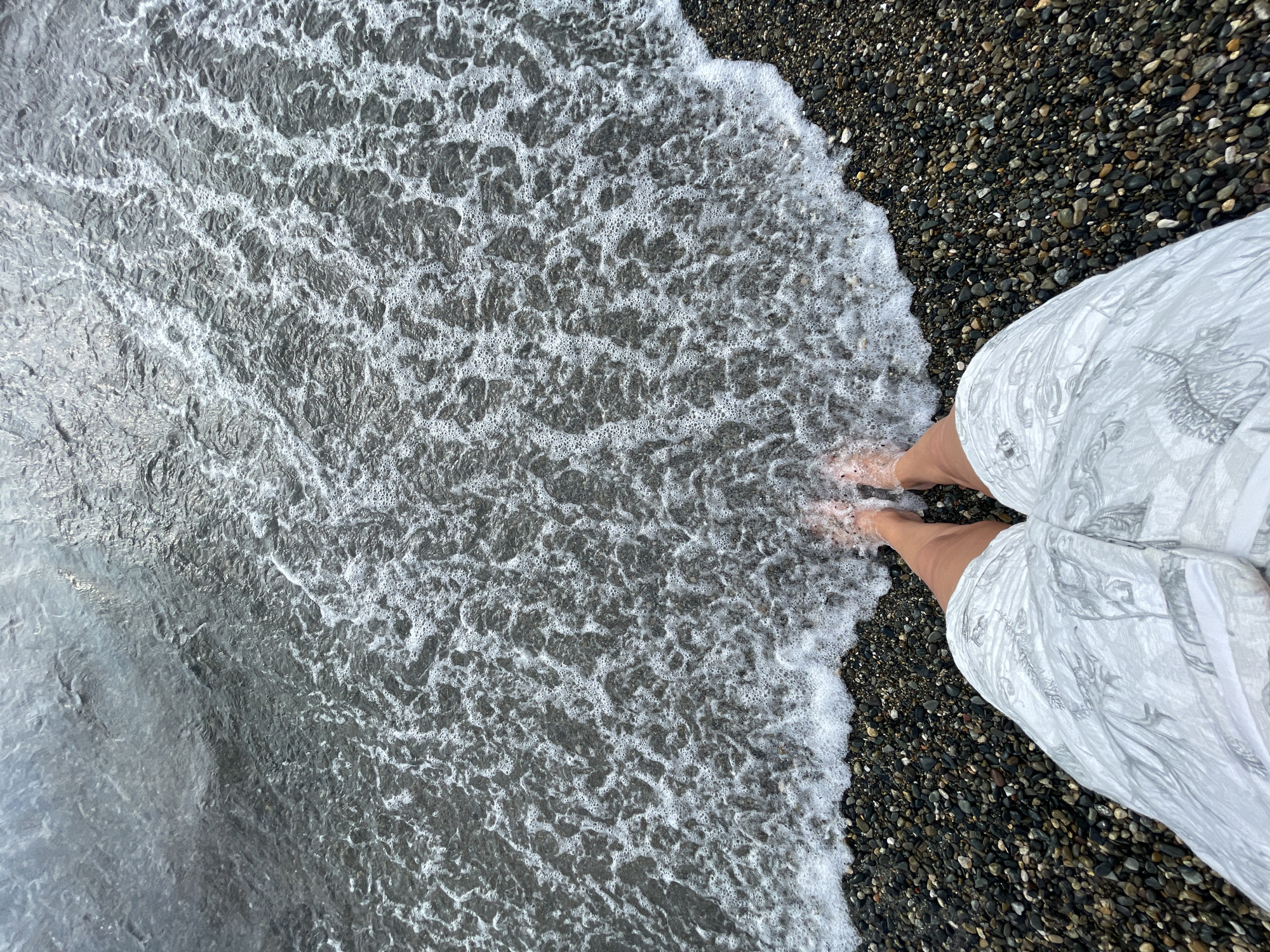 This is a high-angle shot of a person's bare feet standing on a pebbly beach, with the gentle surf washing over them. The water is clear and filled with small bubbles as it recedes, revealing the dark, wet pebbles beneath. The person is wearing a light-colored, patterned garment that drapes over their legs, obscuring the rest of their body. The overall scene suggests a relaxed and peaceful moment at the seaside. The location is identified as Sochi, Russia. There is no visible text in the image.