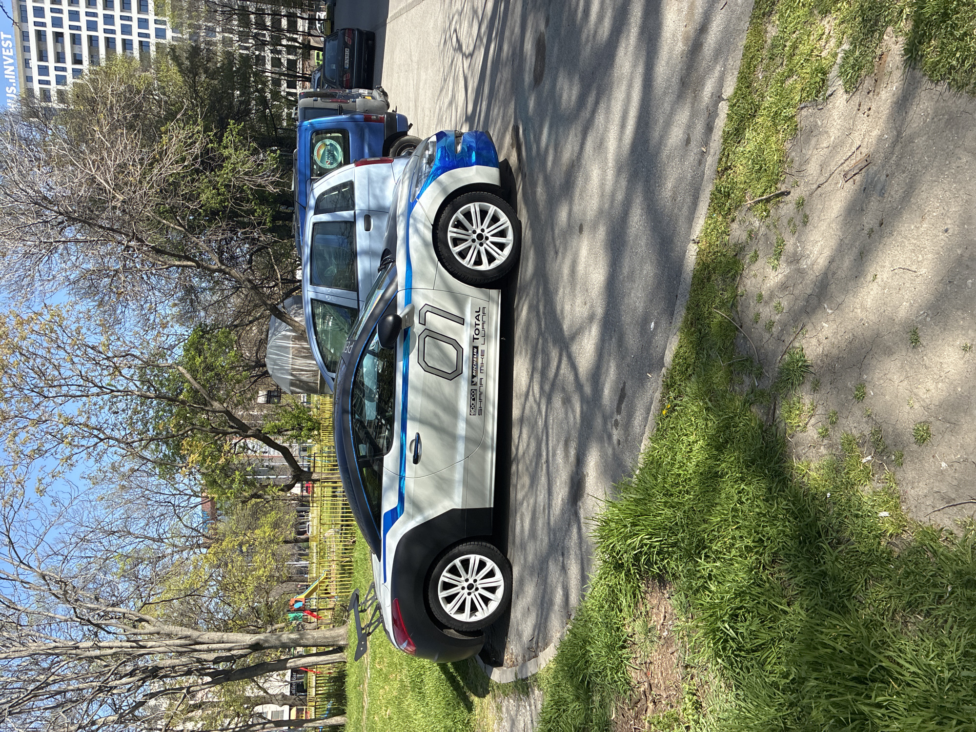 The image shows a white and blue rally car parked on a paved area next to a patch of green grass. The car has the number "01" on its side and sponsors' logos. In the background, there are several other parked cars, a fence, trees, and a tall building with windows. There's a playground nearby, indicating a park or recreational area. The location appears to be an urban environment within Plovdiv, Bulgaria, based on the style of the building and the presence of Cyrillic text in the sponsor's logos. There are no people visible in the shot. Shadows cast by trees indicate daylight.