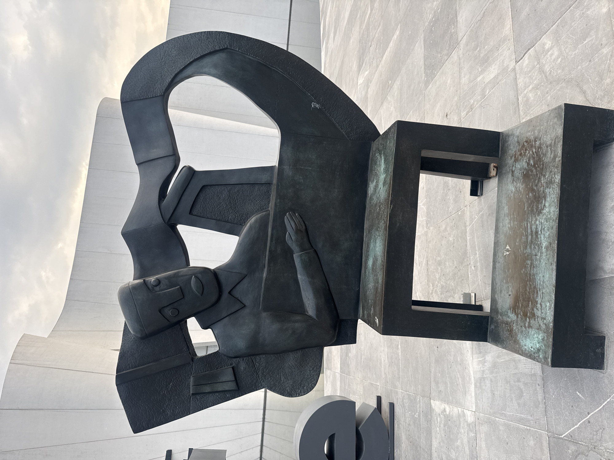 A bronze-colored sculpture of a man is mounted on a grey stone wall. The man, dressed in a suit, is kneeling with his hand raised to his forehead. The sculpture is composed of geometric shapes, including a square frame that connects to the man's head. The sculpture is located in San Bernardino Tlaxcalancingo, Mexico.