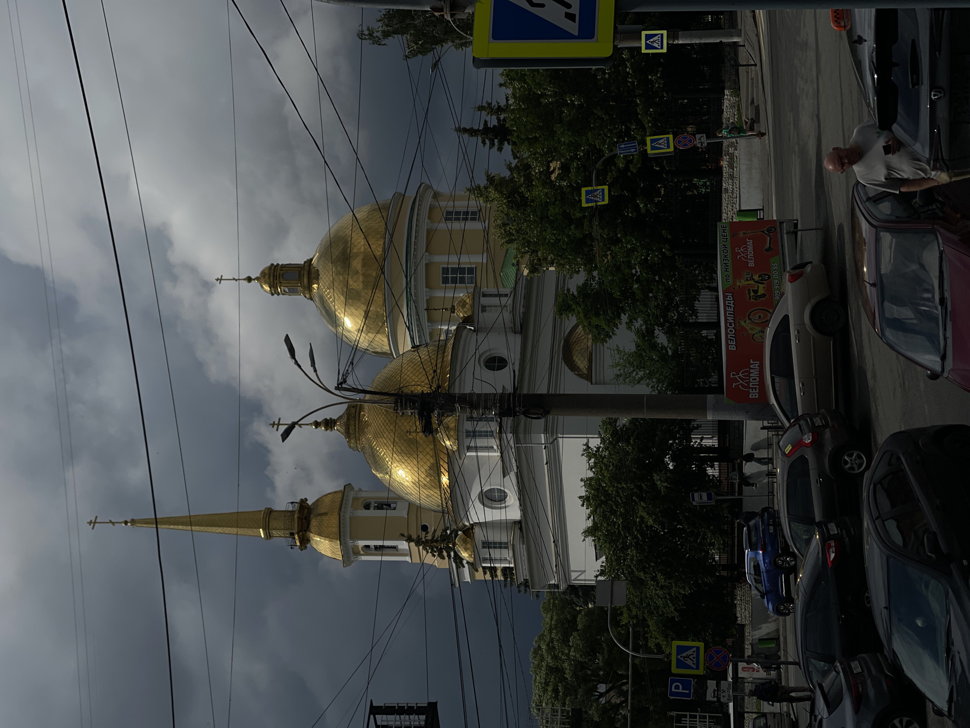 The image depicts a street scene in an urban environment, likely in Russia, given the signage. A prominent Orthodox church with golden domes and a spire dominates the background. The church is a white, multi-story structure with arched windows. In the foreground, a busy street is visible with several cars parked and driving. Traffic lights, street signs, and overhead wires are present, indicating an active urban intersection. Lush green trees partially obscure the lower sections of the church. A billboard advertising bicycles is also visible on the right side of the street. A man in a grey t-shirt is standing near a parked car, looking at his phone. The sky is overcast with dark, heavy clouds, suggesting a potential for rain.