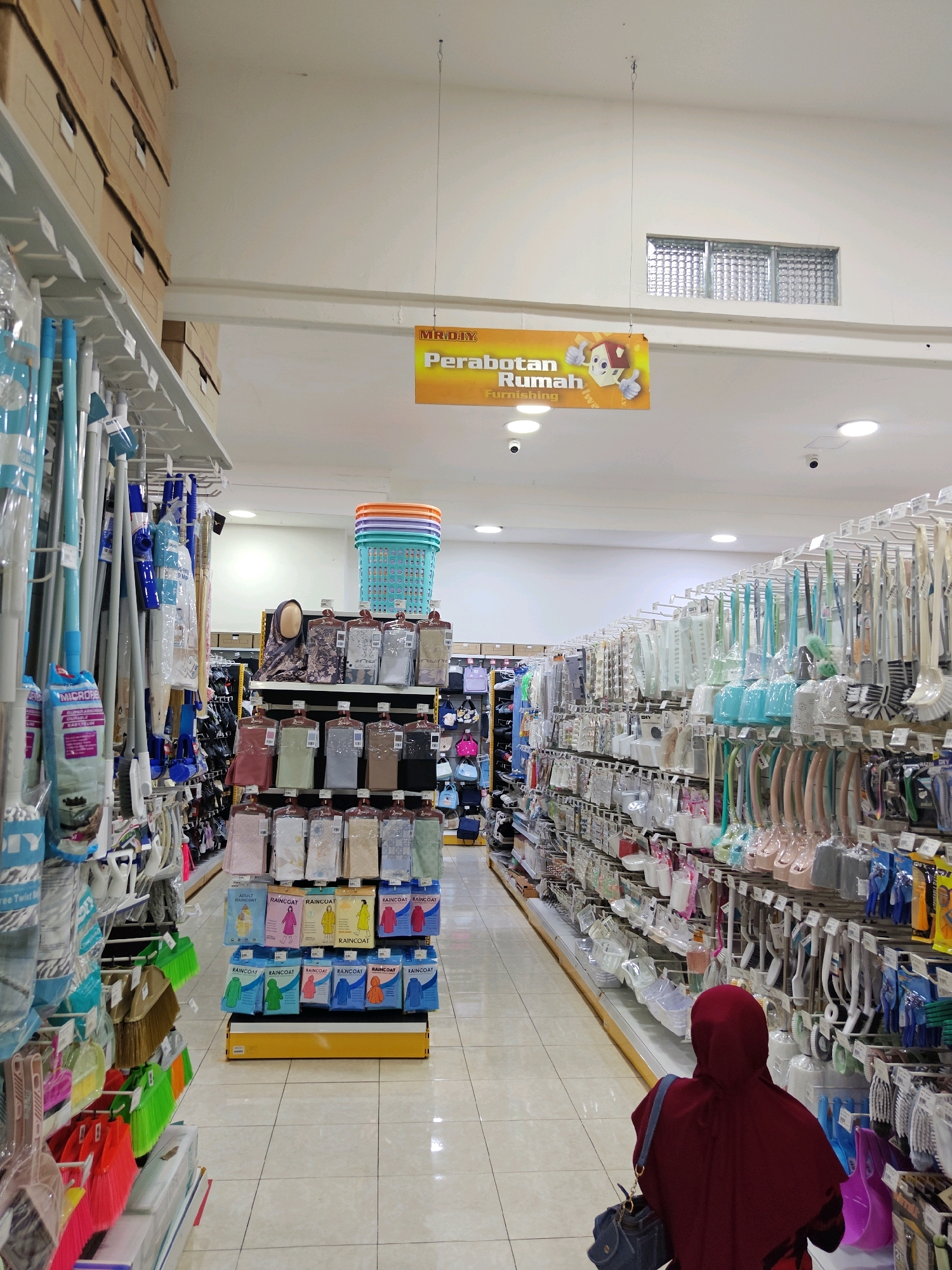 The image shows the interior of a MR.DIY store in Situbondo, Indonesia. The store sells a variety of household goods, including cleaning supplies, kitchenware, and home furnishings. In the foreground, a person wearing a maroon hijab and a blue shoulder bag is browsing the aisles. The aisles are lined with shelves stocked with products. The store appears to be well-lit and clean, with a tiled floor. A sign in the background reads "MR.DIY PERABOTAN RUMAH FURNISHING".