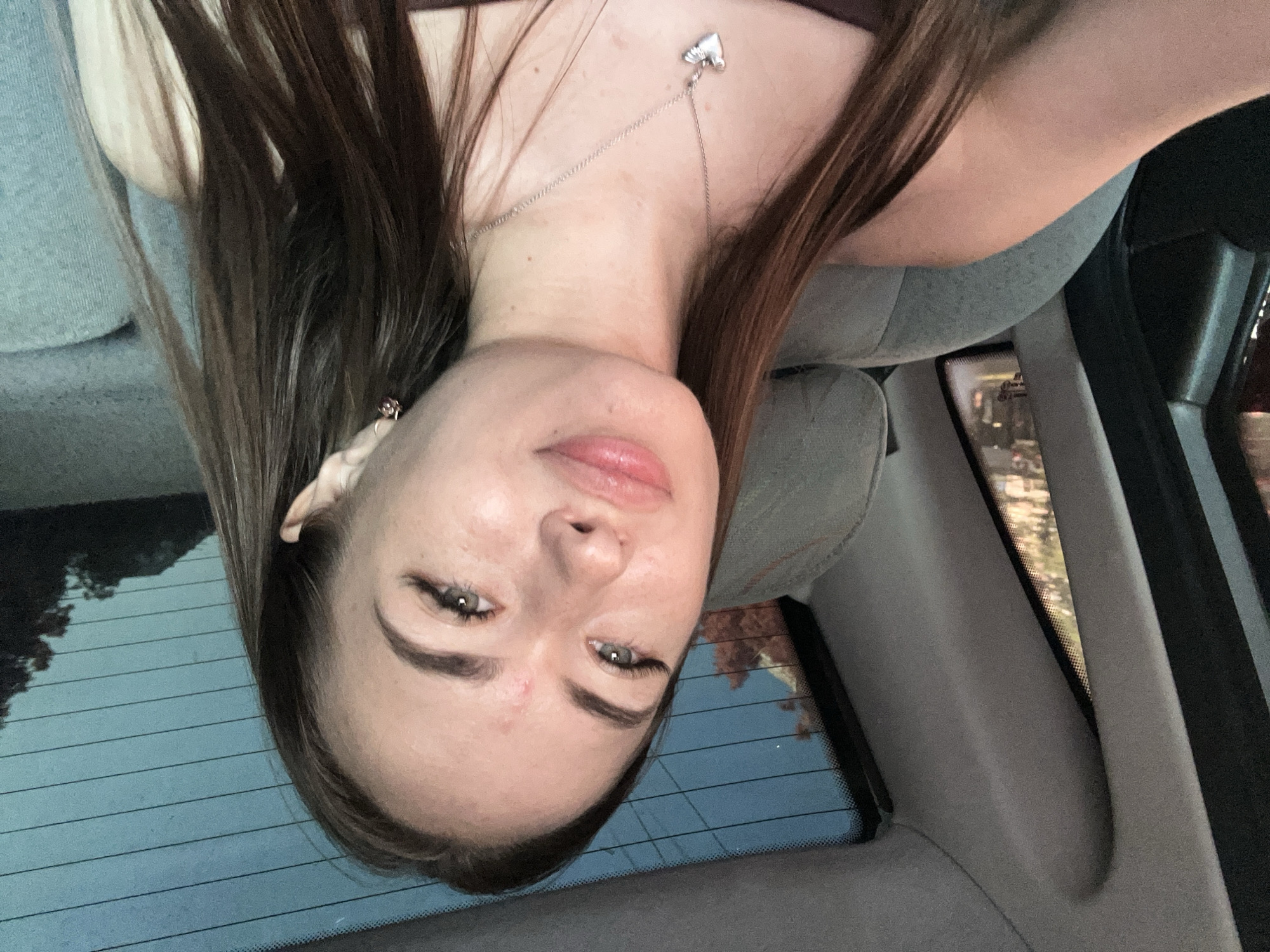 A young woman with long brown hair and green eyes is sitting in the back seat of a car. She is wearing a dark top and a silver necklace with a heart-shaped pendant. The background shows the rear window of the car, which has horizontal lines and a view of trees and the sky. The interior of the car is visible, with gray upholstery on the seats. The lighting suggests it may be evening or nighttime, as there are lights visible through the rear window.
