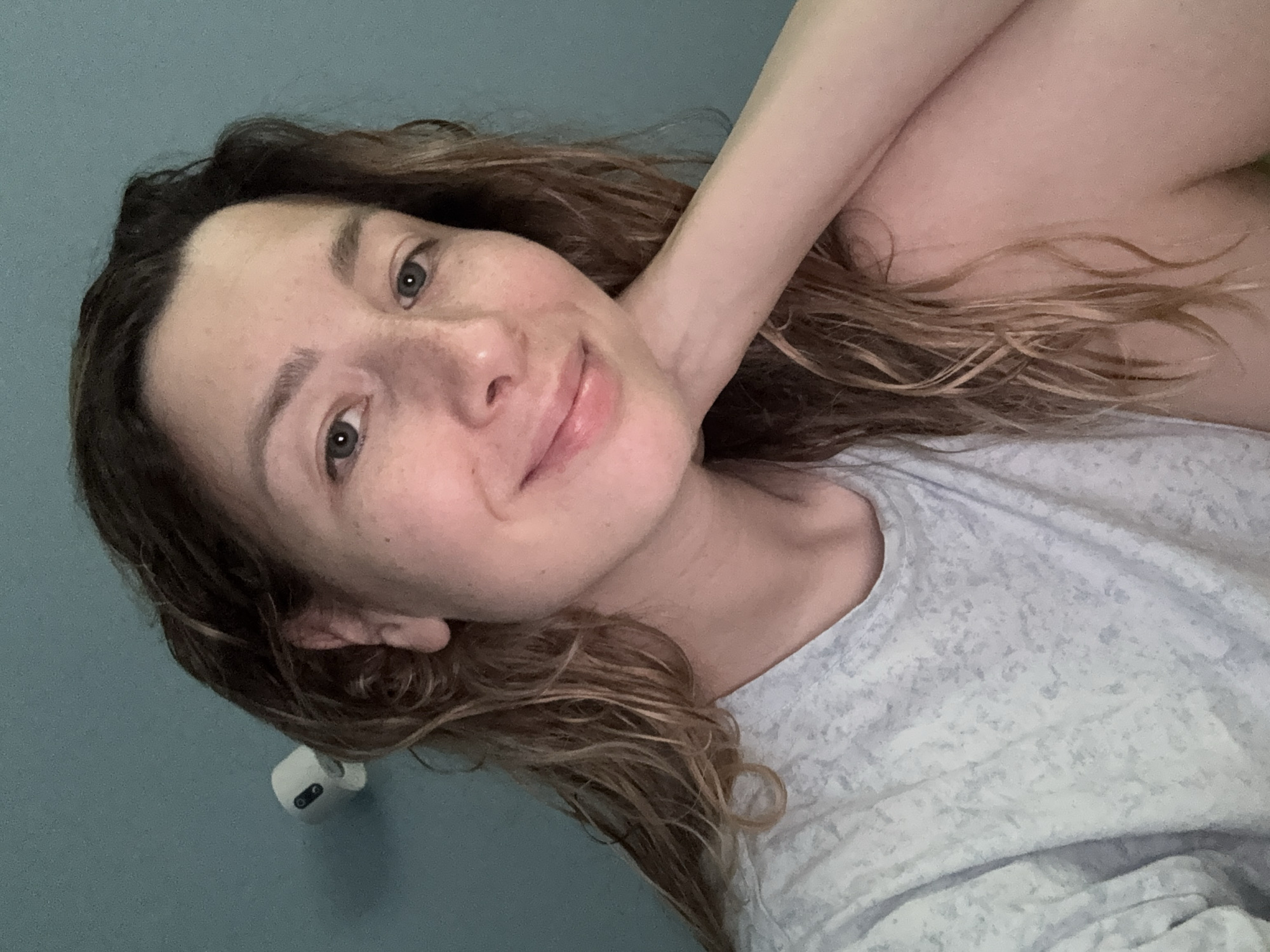 A medium shot image shows a woman indoors, with a blue wall in the background. She has medium-length, wavy, light brown hair and fair skin with freckles. She is wearing a light gray t-shirt with a subtle pattern. Her left arm is bent, and her hand rests on her chin, supporting her head. She is looking towards the camera with a slight smile. A white object, possibly a security camera, is mounted on the wall to the left of her head. The lighting suggests an indoor setting, with soft illumination. The location is not specified as Moscow, Russia.
