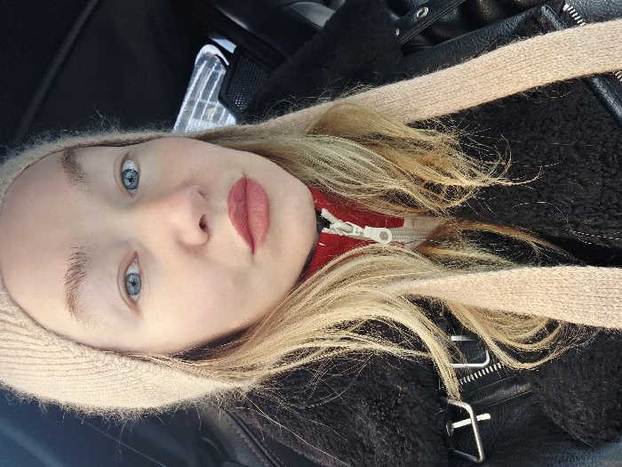 A fair-skinned person with light blonde hair and blue eyes looks at the camera. The person wears a light beige knit hat. A red garment with a white zipper is visible underneath a black jacket that has silver hardware details. The background appears to be the interior of a vehicle.