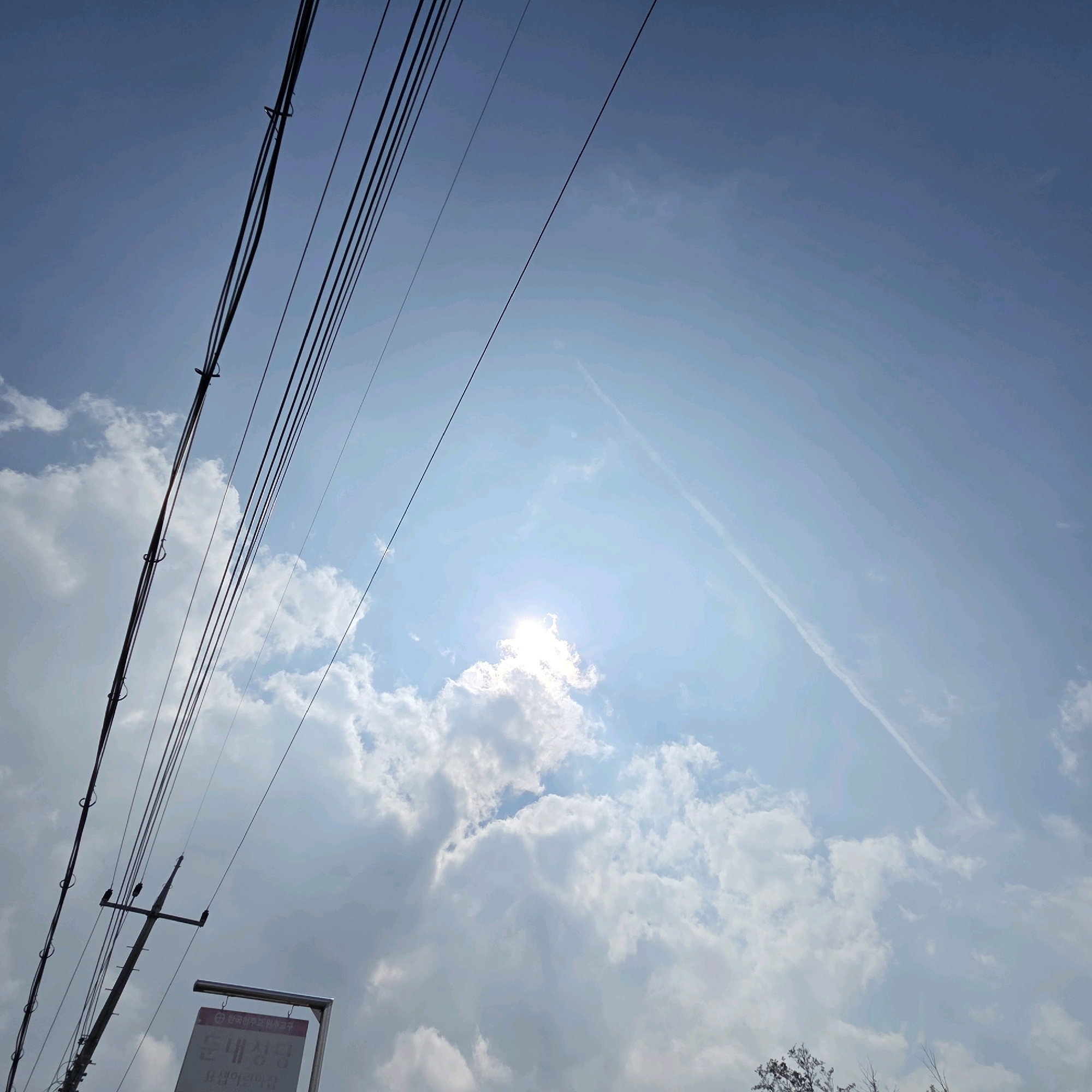 A low-angle shot looks up at a bright, cloudy sky with power lines in the foreground. The sun is visible through a break in the clouds, creating a lens flare effect. A faint contrail from an aircraft stretches diagonally across the sky. In the lower left corner, a portion of a pole with insulators and hanging power lines is visible, along with a sign with Korean text. This scene is located in Dunnae-myeon, South Korea.