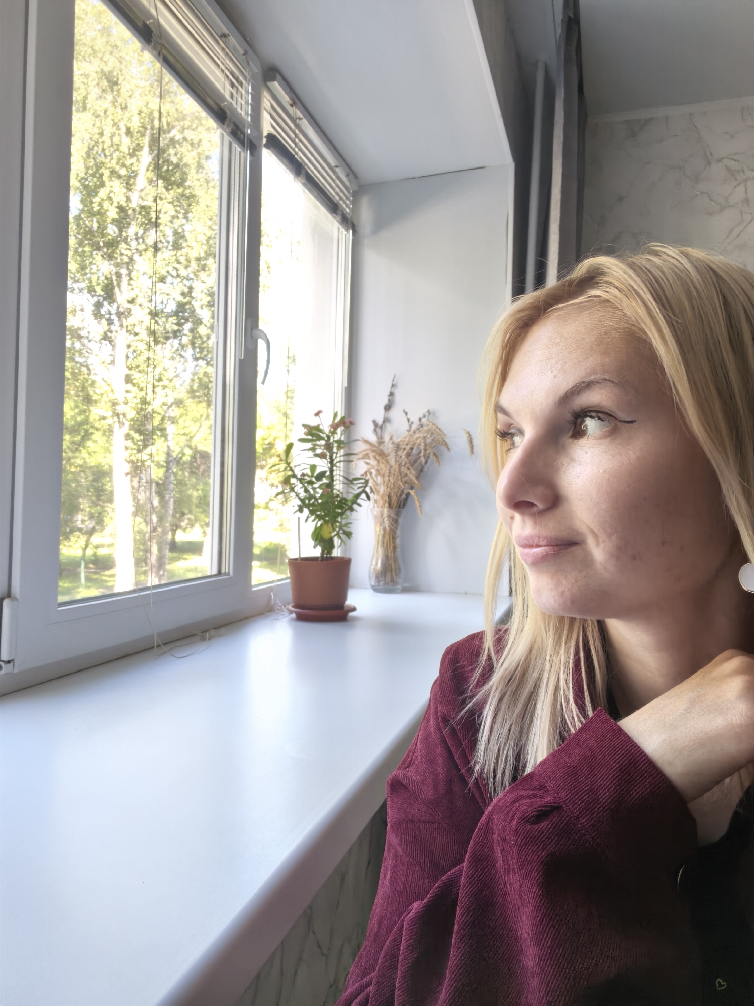 A medium shot captures a blonde woman looking out a window, her face illuminated by natural light. She is positioned on the right side of the frame, with her head turned towards the left. Her hair is styled loosely around her shoulders, and she wears a dark red corduroy jacket. Her gaze is directed slightly downward, and her expression appears contemplative.
In the background, a window offers a view of trees and greenery, suggesting an outdoor setting. A small potted plant sits on the windowsill to the left of the woman, and a vase with dried flowers is visible further to the left. The interior appears to be a room with a white windowsill. The overall scene conveys a sense of quiet observation and introspection. The image is a photograph taken indoors, likely in a residential or office setting. The location of Sterlitamak, Russia, is mentioned in the prompt but is not visually discernable within the image itself.