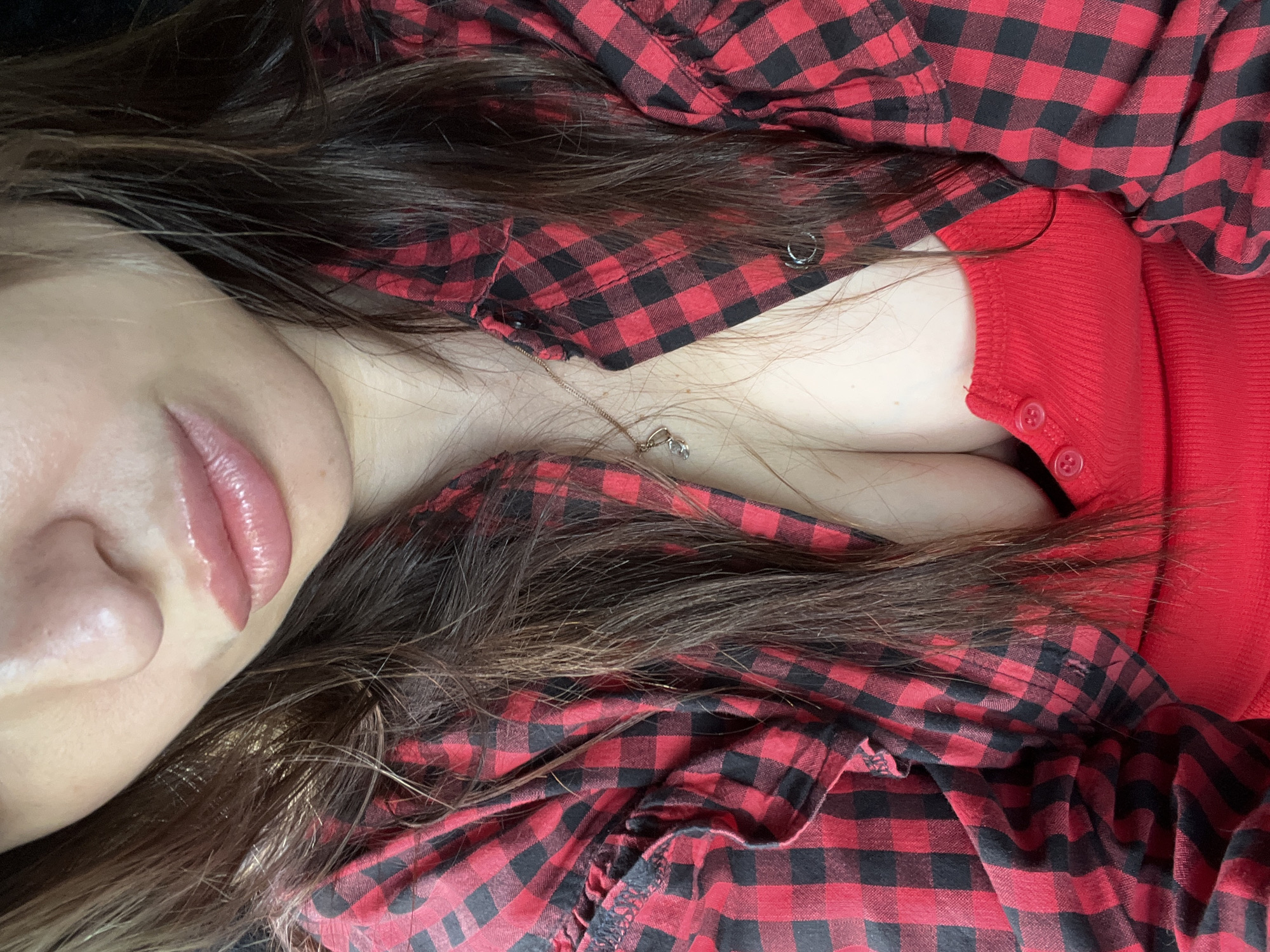 A close-up, chest-up portrait depicts an individual with long, dark brown hair cascading over their shoulders and chest. The visible portion of the face, from the nose down, shows full, glossy, light pink lips. The individual wears an open red and black buffalo check plaid shirt, featuring ruffled details on the sleeves, over a solid red, ribbed knit top. The red top has a three-button placket at the center front, revealing cleavage. A thin, gold-colored chain necklace with a small, light-colored charm rests on the décolletage. The background is dark and featureless, providing no environmental context. The location is Unknown, Unknown.