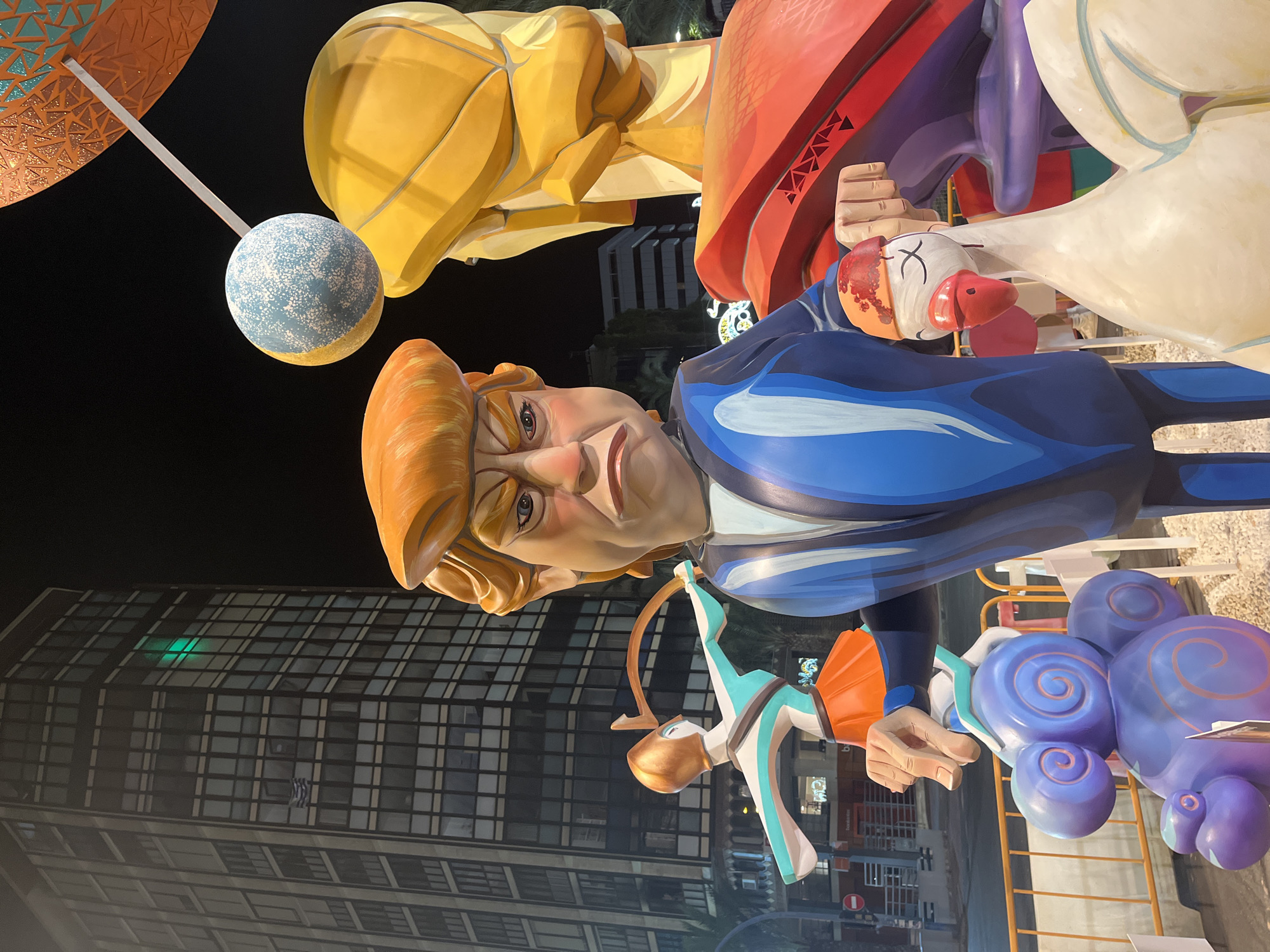 The image displays a nighttime scene featuring large, colorful effigies, likely part of a festival, with a modern high-rise building in the background. In the foreground, a caricature of Donald Trump is depicted wearing a blue suit, with an exaggerated angry expression. To his right, a large yellow effigy with a rounded head is visible. In the middle ground, a figure resembling an archer, with a golden helmet and teal and orange attire, is posed. Also in the foreground, a white goose-like effigy with a bandaged head and red beak stands prominently. The effigies are placed in an outdoor urban environment, with streetlights and a building illuminated behind them. The architecture suggests a contemporary cityscape. The presence of what appears to be a small Greek flag on the building implies a possible connection to a specific event or location, but the scene is predominantly characterized by the large-scale satirical figures. The image was captured in Alicante/Alacant, Spain, likely during the Fallas festival, which features similar effigies.