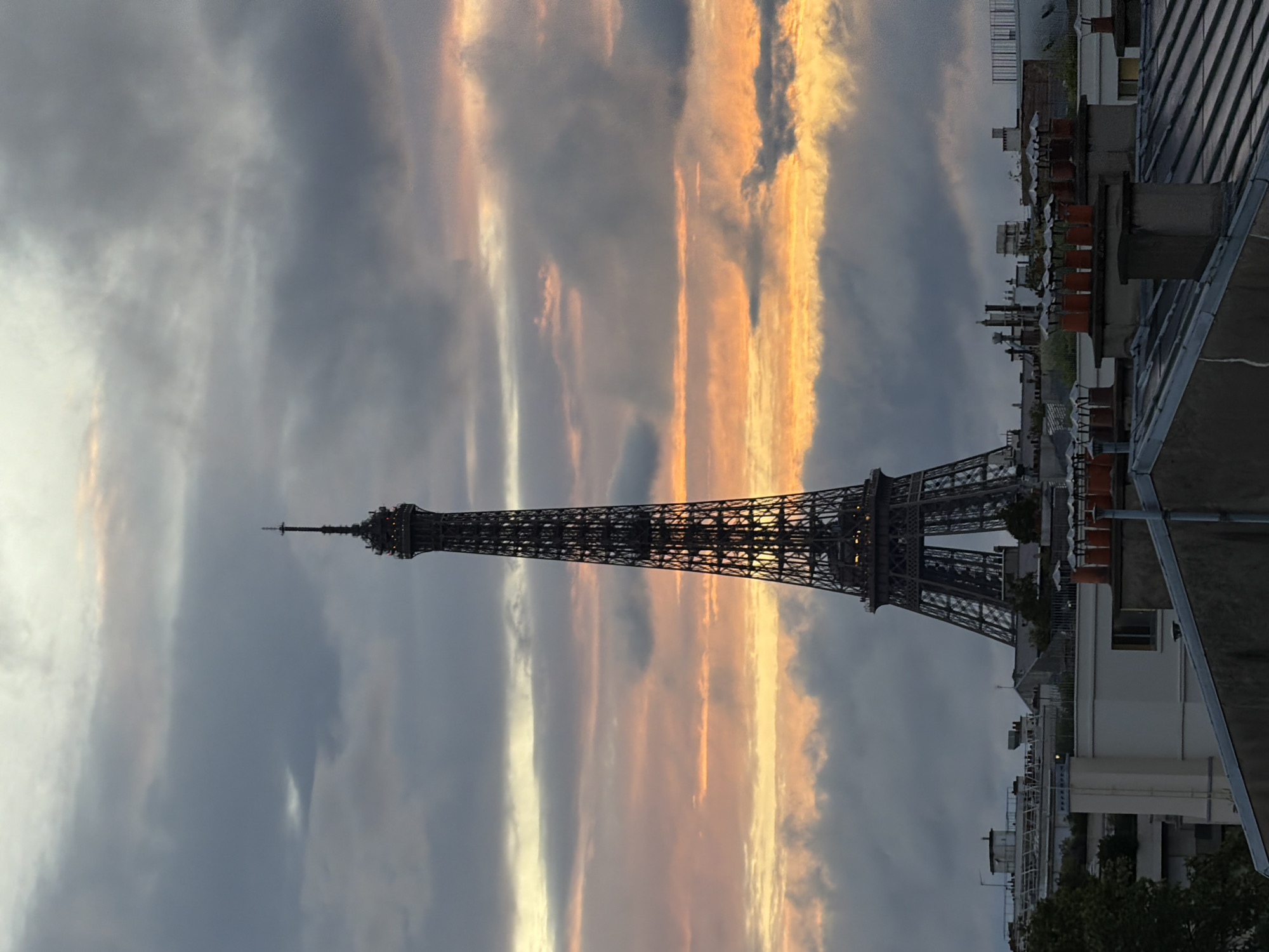 This rotated photograph captures the iconic Eiffel Tower against a dramatic sunset or twilight sky in Paris, France. The towering structure of the Eiffel Tower, rendered in a dark silhouette, stands prominently slightly left of the image's center, its intricate lattice work discernible.
The sky is a dominant feature, filled with a dynamic interplay of clouds. On the left and behind the tower, the clouds are predominantly shades of grey and muted blue. However, to the right and sweeping across the mid-section of the sky, brilliant streaks of warm orange, yellow, and white light pierce through and illuminate other clouds. These elongated, luminous formations create a striking contrast with the darker clouds, suggesting either the sun setting or rising just out of frame.
In the foreground, occupying the lower right portion of the image, are the rooftops of traditional Parisian buildings. These are depicted in shadow, revealing architectural details such as numerous chimneys, ventilation systems, and characteristic slanted slate roofs. The scene conveys a majestic and somewhat serene atmosphere, emphasizing the beauty of Parisian architecture and the dramatic natural light of dusk or dawn. There are no people or visible text in the image.