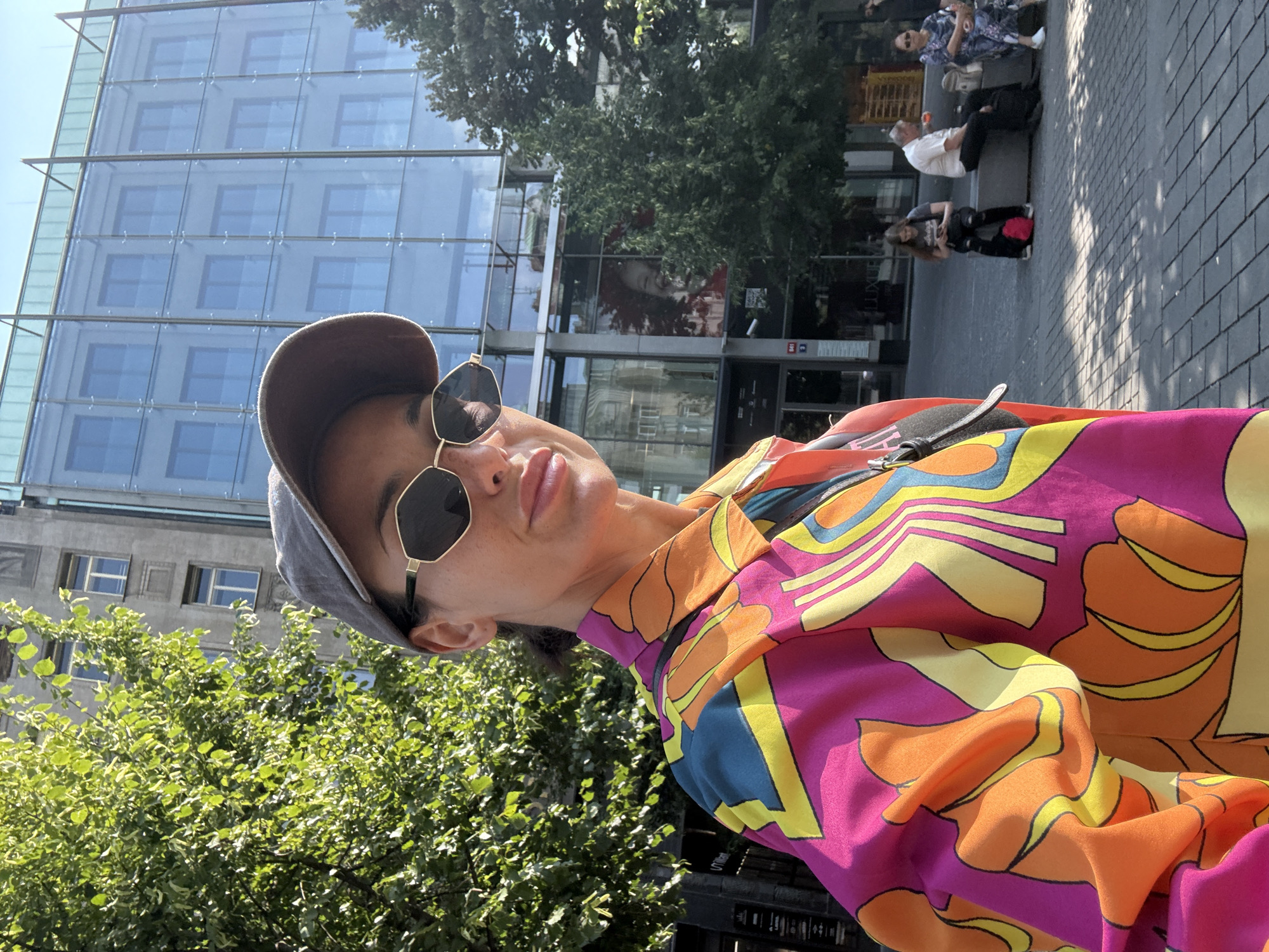 In this medium shot, a woman wearing a colorful, patterned shirt and a baseball cap stands in the foreground, looking towards the camera. Her sunglasses obscure her eyes, but her lips are slightly parted. Behind her, a modern glass-fronted building reflects the sky and surrounding trees. To the left, a lush green tree with sun-dappled leaves occupies a significant portion of the frame. In the middle ground, several people are seated on a bench, including a woman looking at her phone and an older man. The ground is paved with grey square tiles. The scene appears to be an outdoor public space in Prague, Czechia, possibly near a modern commercial area.