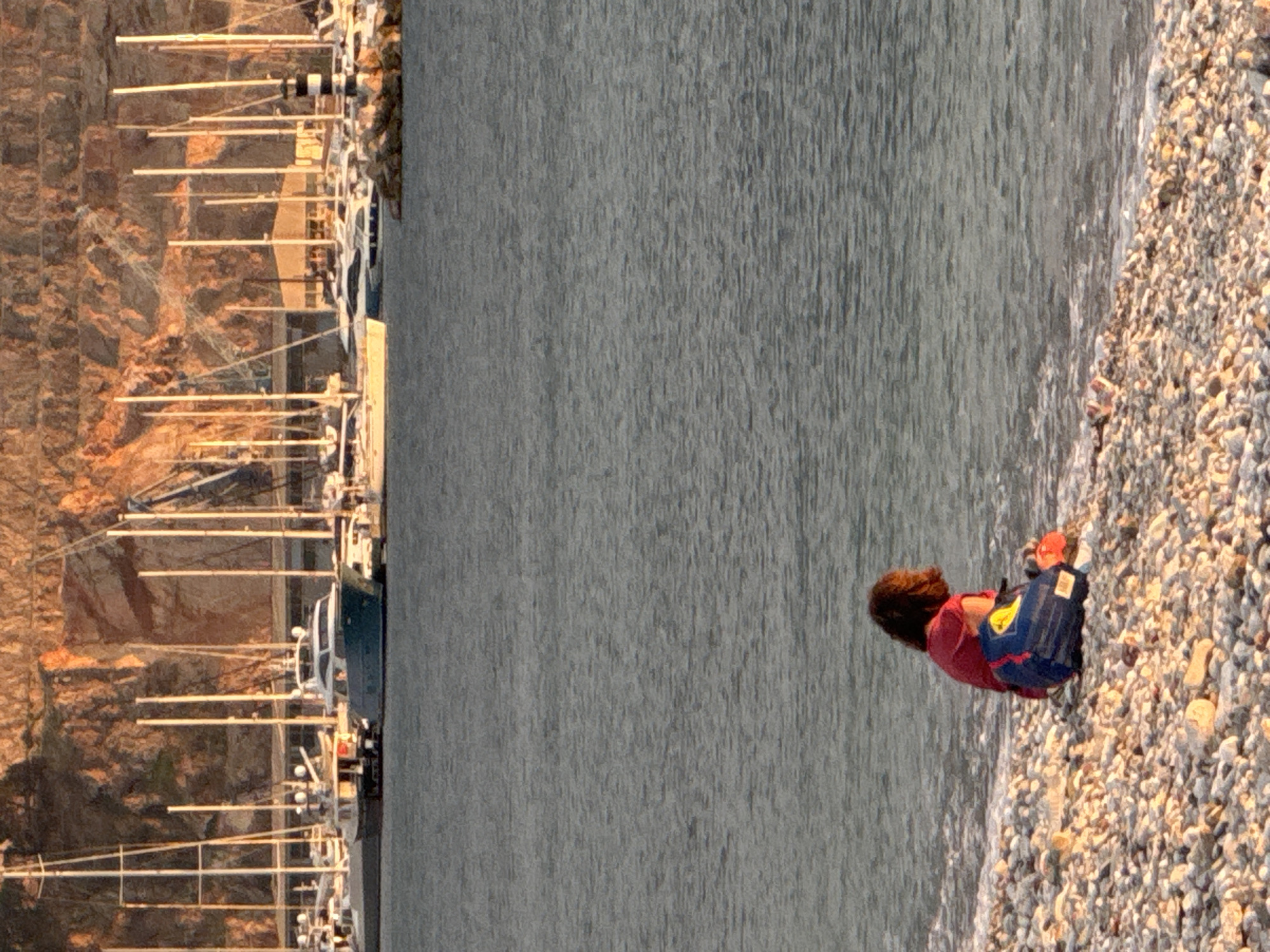 The image depicts a beach scene in Šušanj, Montenegro. A person is kneeling on a pebble beach near the water's edge. They are wearing a reddish top and have a blue and red backpack next to them. The water stretches out towards a harbor filled with numerous boats and yachts docked along a quay. Beyond the quay, a rocky, sunlit cliff face is visible.
