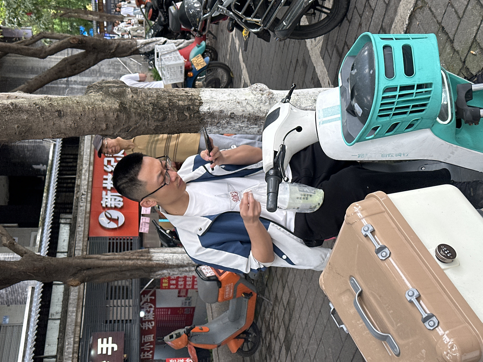 A medium, eye-level shot depicts a man sitting on a scooter and looking at his phone while in Shapingba District, China. The man is wearing glasses and a white t-shirt with a blue and white jacket. He is sitting on a white scooter with a teal front basket and a light brown suitcase next to him.
In the background, there are several other scooters parked and a building with a red sign that reads "重庆 老麻抄手" (Chongqing La Ma Chao Shou), which translates to "Chongqing Spicy Noodle Shop". There is also another sign that reads "手 庆小面 老麻抄手" (hand Qing Xiao Mian La Ma Chao Shou). A tree trunk and branches obscure some of the background. The street is paved with light gray bricks.