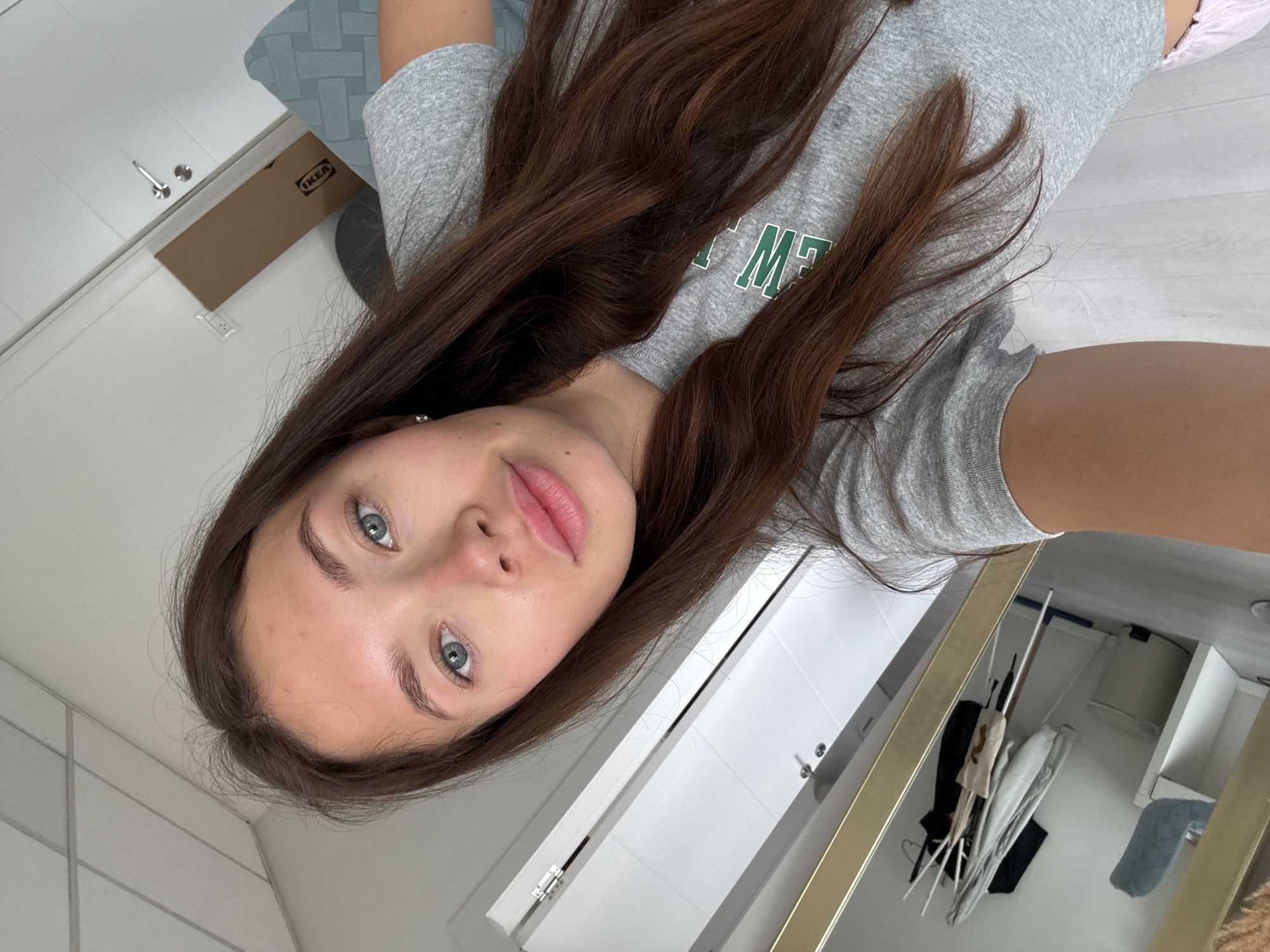 The image is a selfie taken indoors, likely in a bathroom. A young woman with brown hair and blue eyes is the primary subject. She is wearing a gray t-shirt with green lettering. Her skin appears fair. The background reveals a white tiled wall, a white cabinet with chrome handles, and a gold-framed mirror reflecting various items, including clothing and toiletries. Additionally, there is an IKEA box on a shelf. Without further information, the location within Ramat Gan, Israel, cannot be determined.