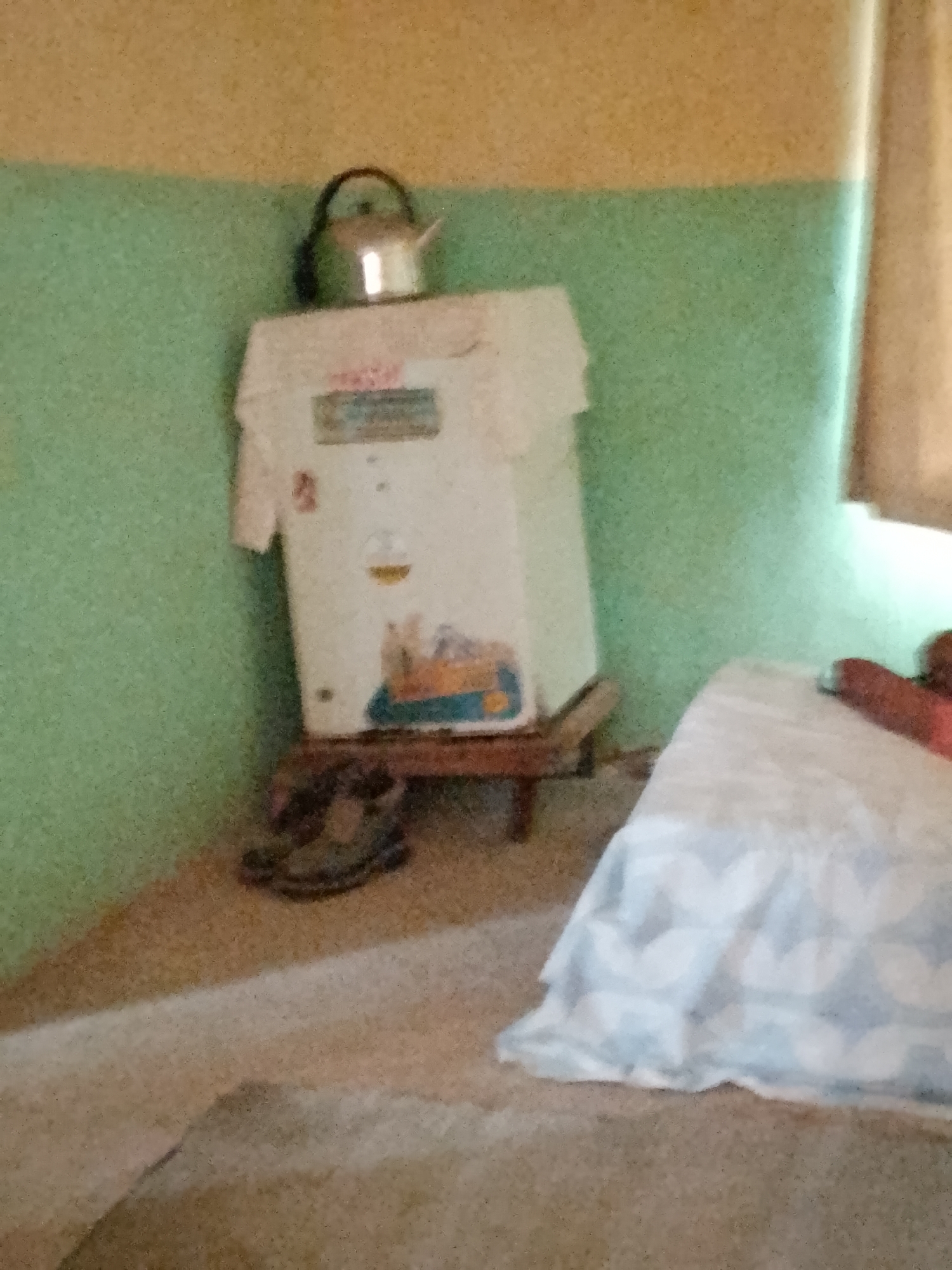 In an indoor scene in a room with green and yellow walls, a white appliance, possibly a refrigerator, stands on a wooden base. A silver kettle with a black handle is placed on top of the appliance. To the left of the appliance, a pair of dark-colored shoes are visible. A bed with a white and blue patterned cover is present on the right side of the image. A reddish-brown object, potentially a pillow or blanket, is on the bed. The floor is covered in a light-colored carpet.