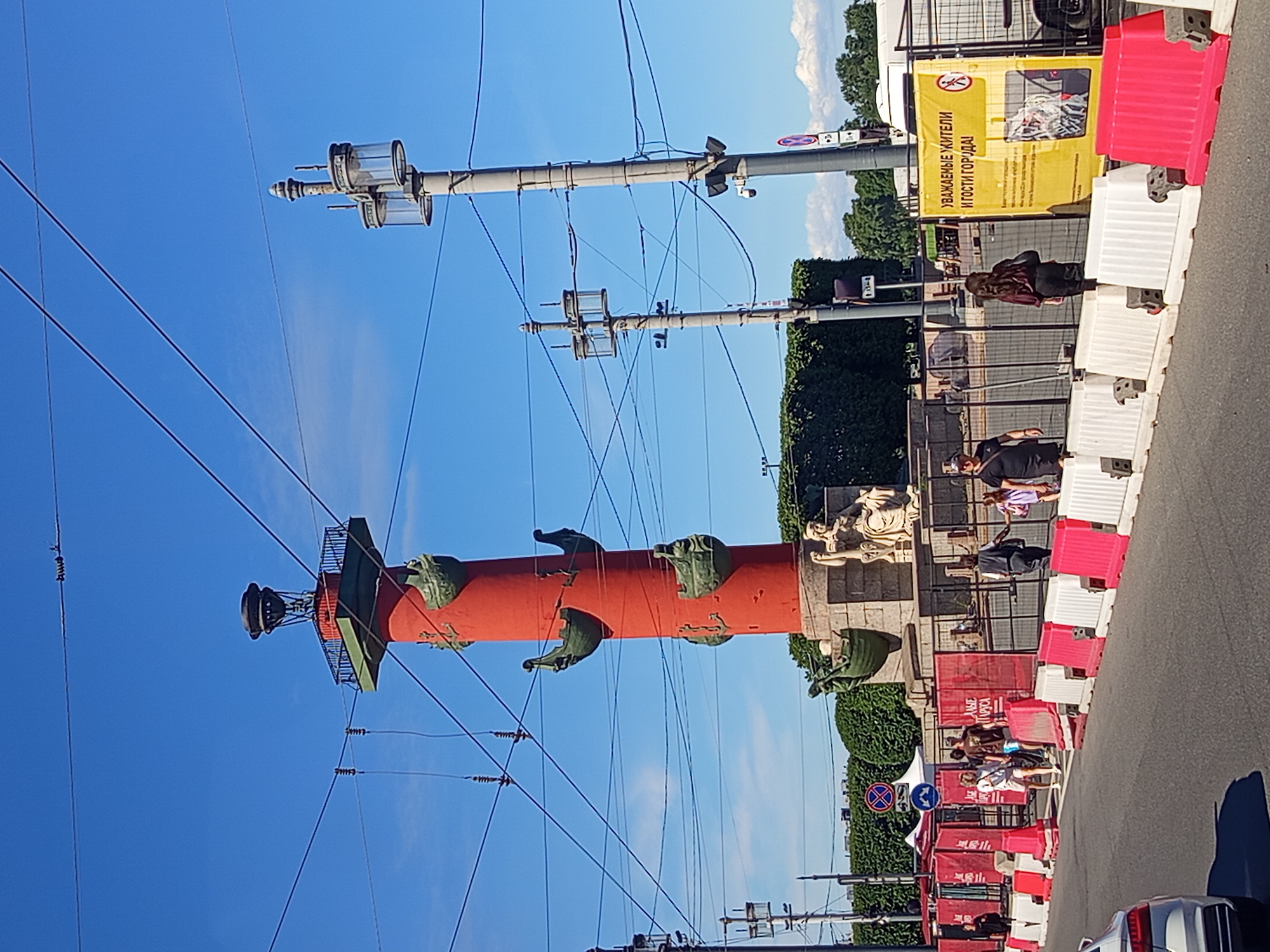 The image captures a street scene dominated by a tall, red Rostral Column with ship prows adorning its sides. Two lampposts stand to the right of the column, and several power lines crisscross the clear blue sky. In the foreground, a road is lined with white and pink plastic barriers. A white car is partially visible on the left. People are walking on the sidewalk behind the barriers, some near a construction fence. A large yellow sign with Russian text is affixed to a fence on the right side of the image. The background features dense green foliage and a white tent.