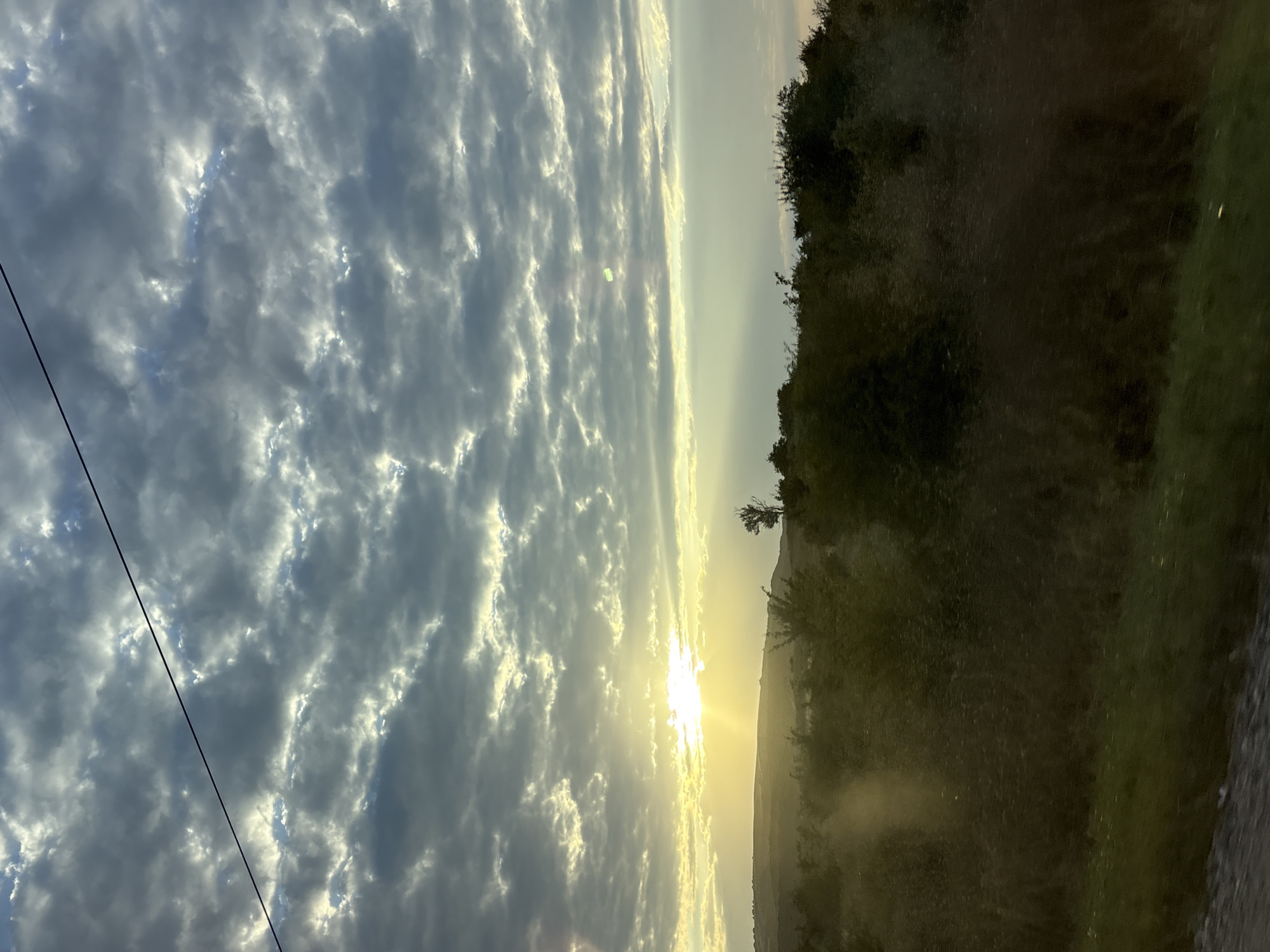 The image shows a scenic landscape with a dramatic sky over rolling hills and vegetation. The sun is low on the horizon, casting a warm glow and creating lens flare. The sky is filled with textured clouds, some of which are backlit by the sun, creating a sense of depth and dynamism. A power line stretches diagonally across the upper portion of the image. The foreground consists of dark, dense vegetation, with some lighter green grass visible at the bottom. The overall impression is one of a natural, rural setting, possibly during sunrise or sunset. The location is Lysogorskaya, Russia. There are no people or animals visible in the image.