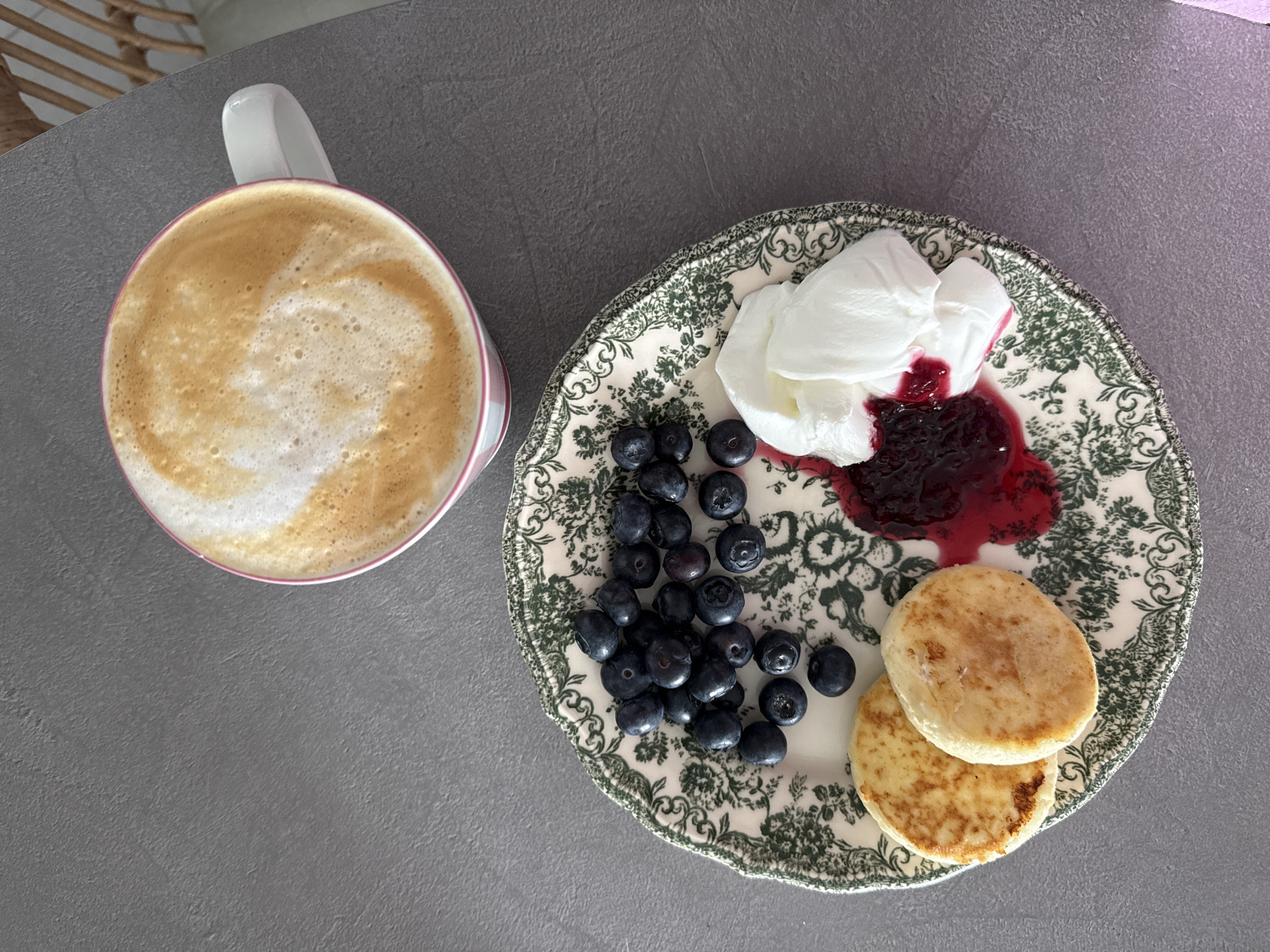 A high-angle shot displays a breakfast scene on a gray surface, likely a table. To the left, a mug filled with a foamy latte rests. Adjacent to the mug is a patterned plate containing a portion of blueberries, a dollop of white cream or yogurt mixed with a dark, possibly berry, jam, and two small, round pancakes or blinis. The scene is indoors, and while there is no specific indication of it being in Tel-Aviv, Israel, no information contradicts this assertion.