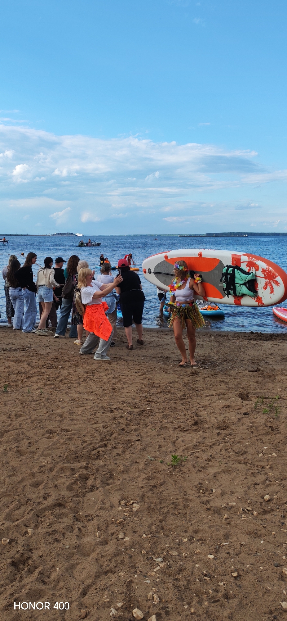 The image depicts a beach scene in Chaikovsky, Russia, with a group of people gathered near the water. In the foreground, several individuals are standing on a sandy beach. A woman in the center right is carrying a paddleboard over her shoulder and is dressed in a hula skirt and a floral lei. Behind her, other people are interacting, with some looking towards the water. In the background, the Kama River is visible with several boats and a large vessel in the distance. The sky is blue with scattered clouds. The overall atmosphere suggests a recreational event or gathering.