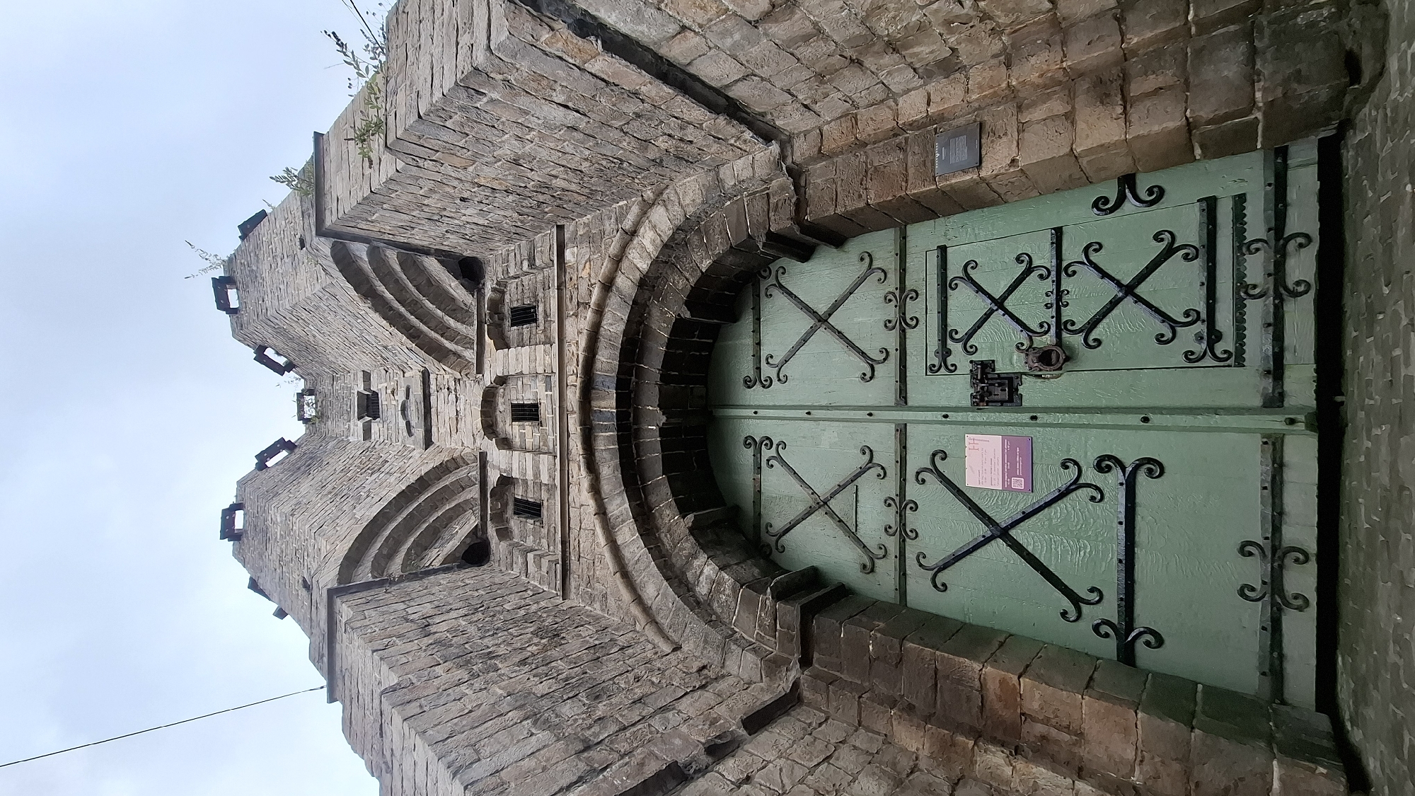 The image shows a low-angle, wide shot of the imposing brick facade of the St. Bavo's Cathedral in Ghent, Belgium, focusing on its large, ornate, mint-green double doors. The doors are decorated with black wrought-iron detailing, forming intricate criss-cross patterns and scrolling filigree. A heavy metal lock and a circular door knocker are visible on the right door. Above the doors, the stonework features multiple arched recesses and small, square windows, suggesting a historical architectural style. The overall structure tapers upwards towards a crenellated top, with glimpses of the sky overhead. The left side of the image shows a thin wire extending from the upper left corner. A small informational plaque is affixed to the stone wall to the right of the doors. The ground in front of the doors appears to be paved with cobblestones.